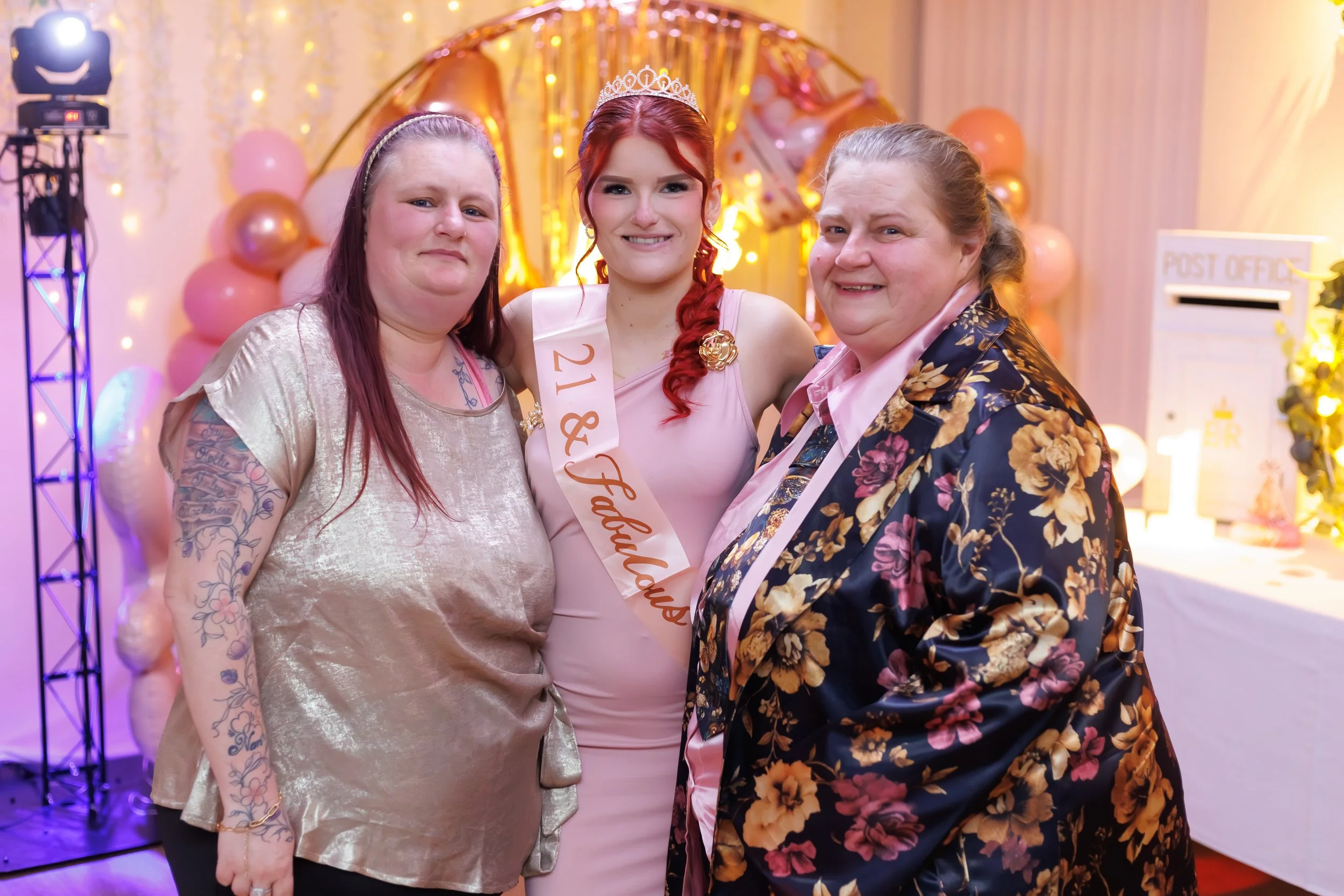 Three women standing together at a celebration, with a red-haired woman in the middle wearing a tiara, sash, and a pink dress. The woman on the left has tattoos on her arm and wears a shiny beige top. The woman on the right has her hair tied back and