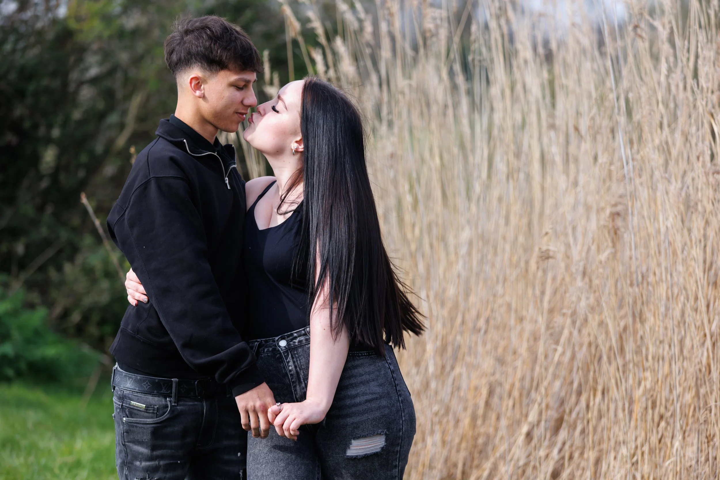 A young man and woman stand close together outdoors, about to kiss, with a field of tall grasses behind them.