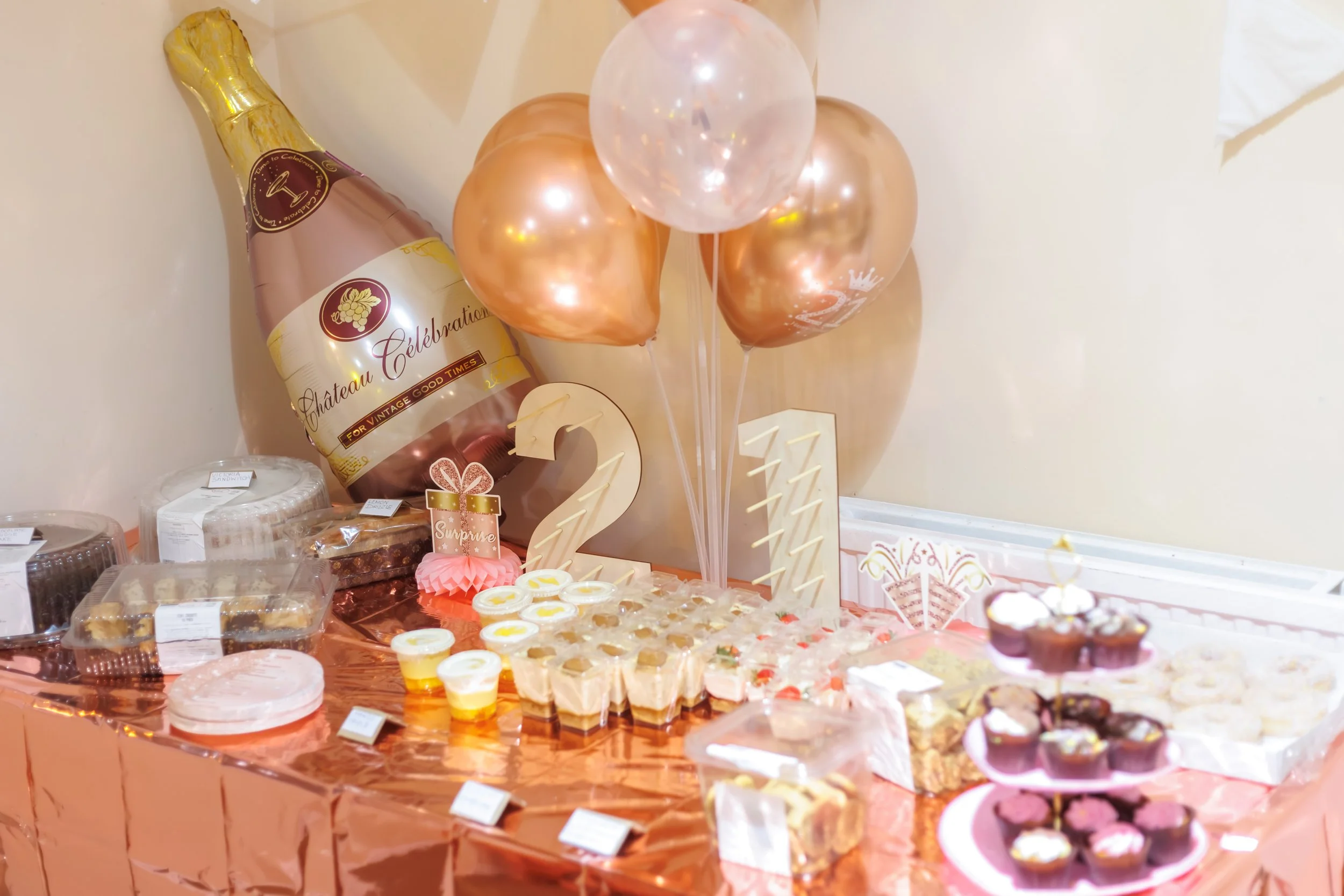Decorations and party treats for a celebration, including a champagne bottle balloon, pink, gold, and white balloons, cupcake desserts, and a variety of sweets on a table.