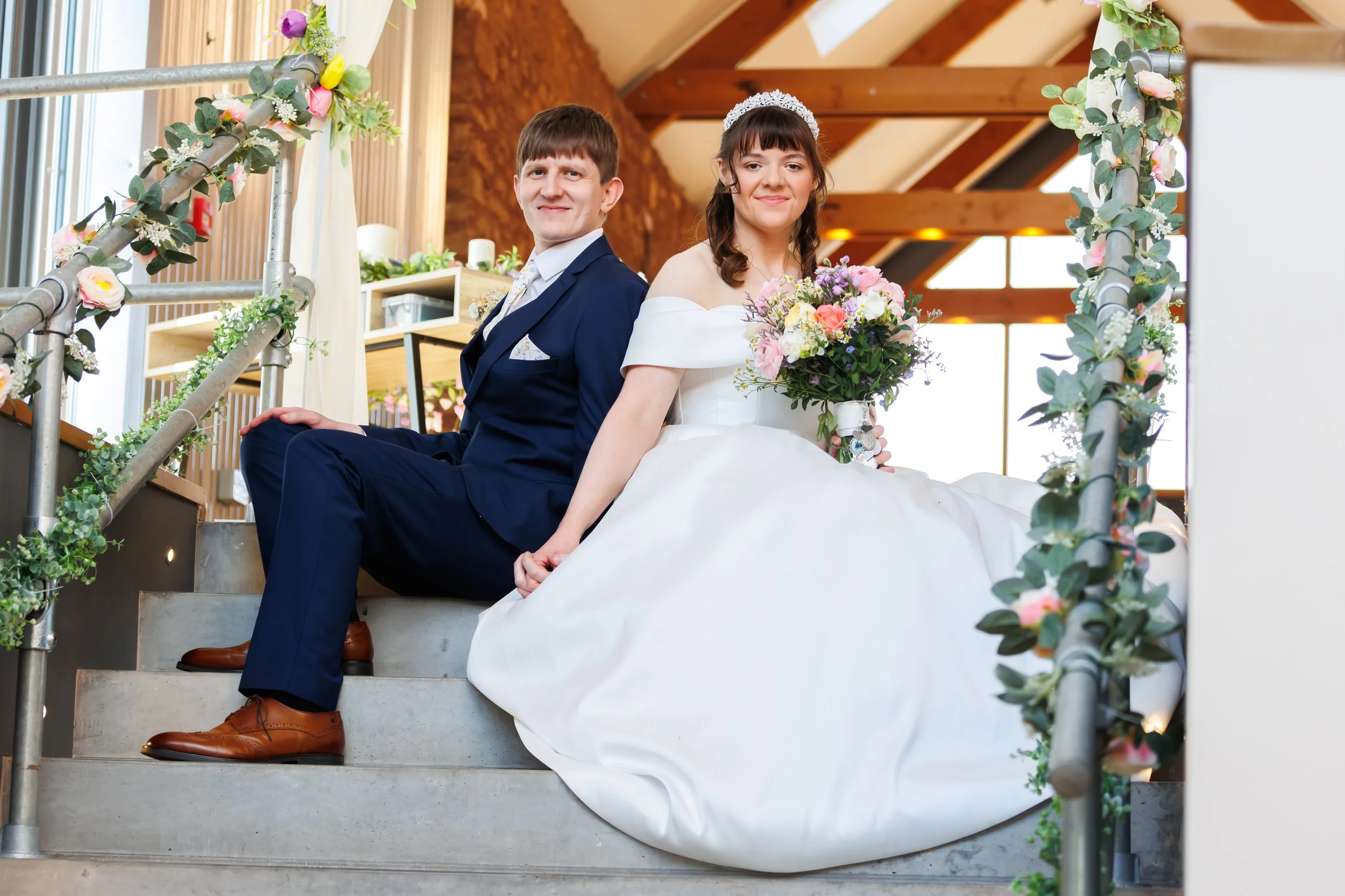 A bride and groom sitting back-to-back on a staircase during their wedding ceremony, surrounded by floral decorations on the railing, with a wooden and natural background.