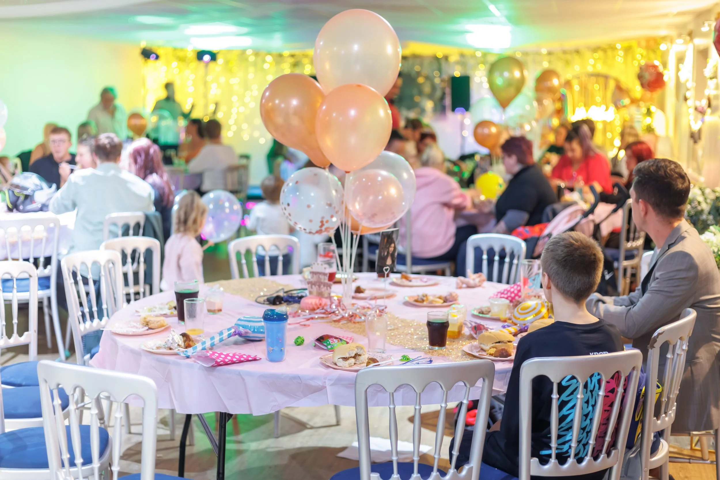 Colorful birthday party with balloons, tables with plates and food, and guests seated indoors with festive decorations.