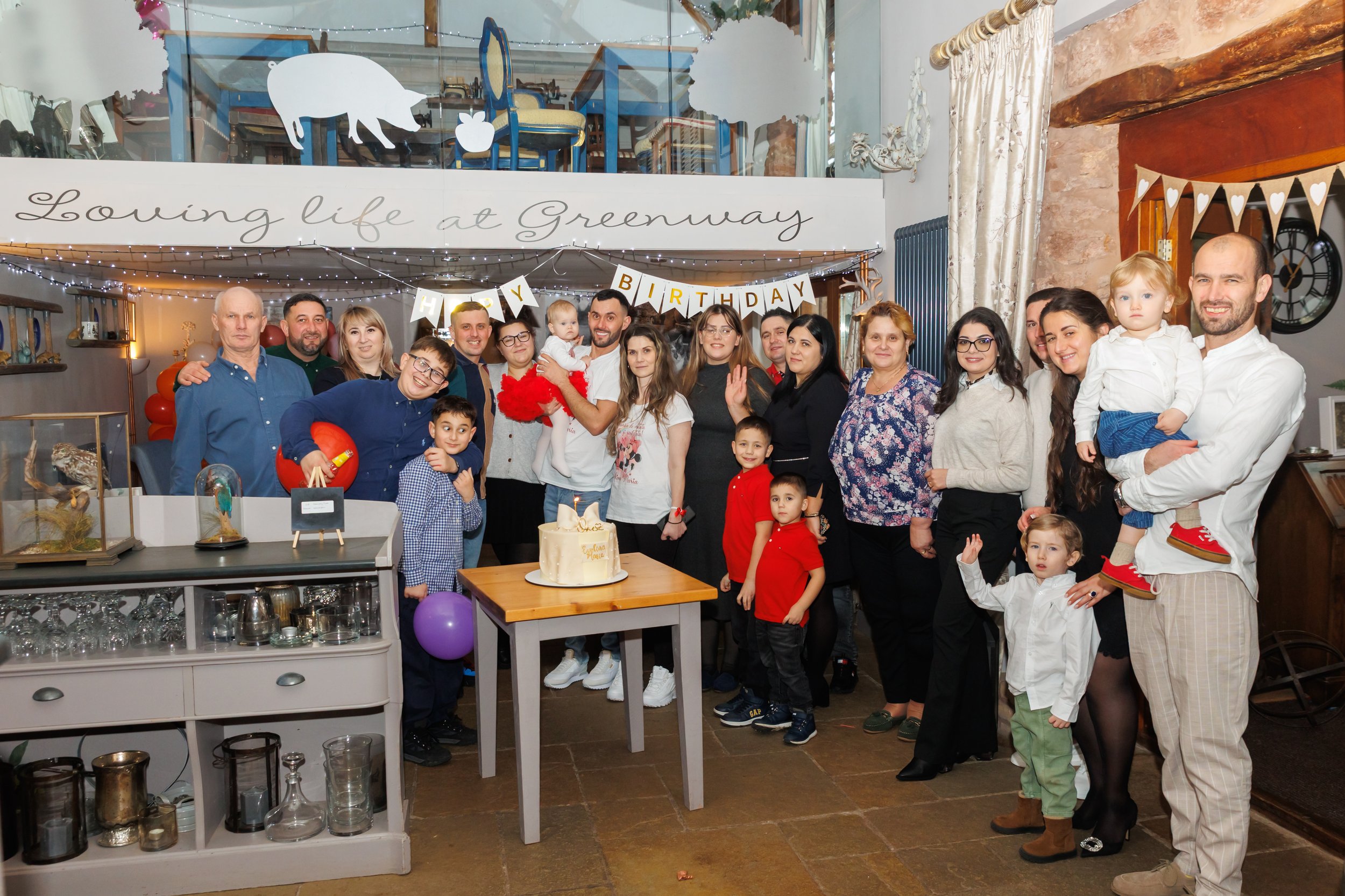 A large group of people, including children, gathered for a birthday celebration in a cozy indoor space with rustic decor. There is a cake on a table in front, and everyone is smiling and posing for the photo. Decorations include a 'HAPPY BIRTHDAY' b
