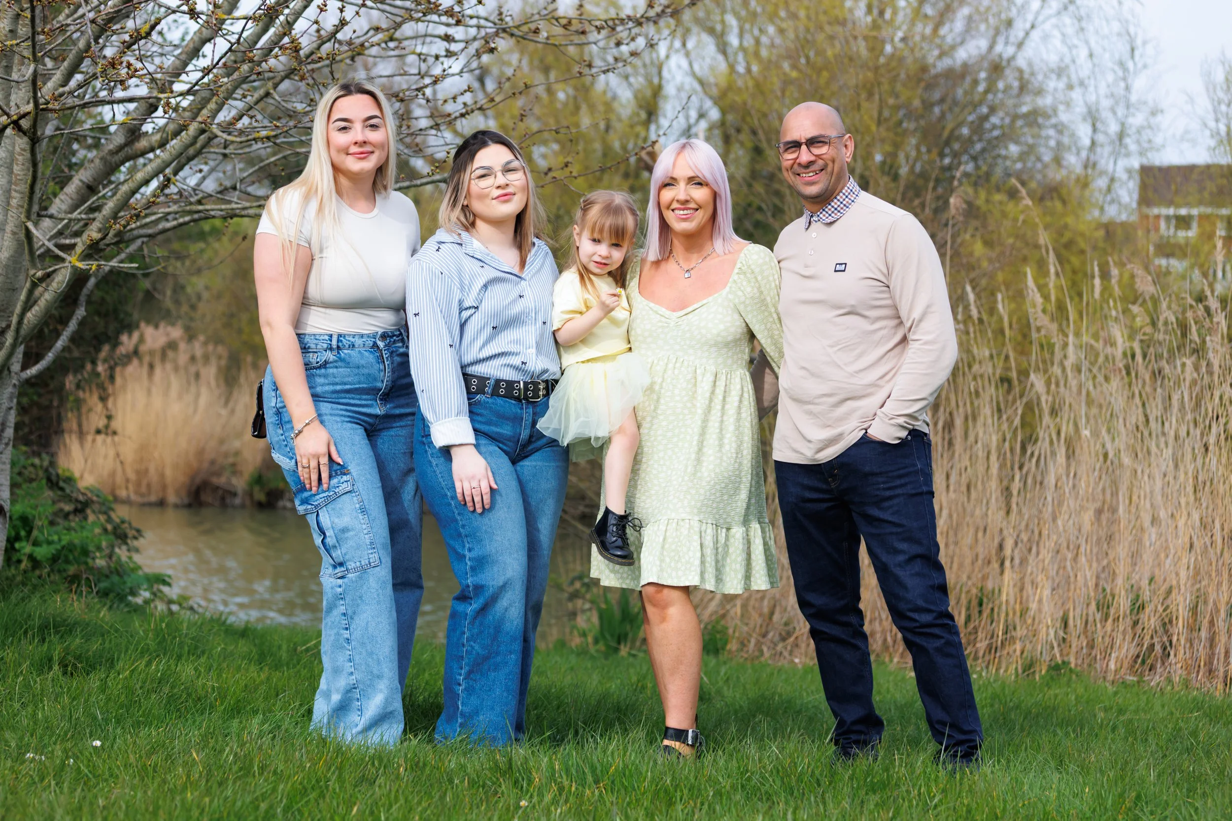 A group of five people, including three women, a young girl, and a man, standing outdoors in a park with trees and a small river behind them. They are smiling and posing together on a grassy area.