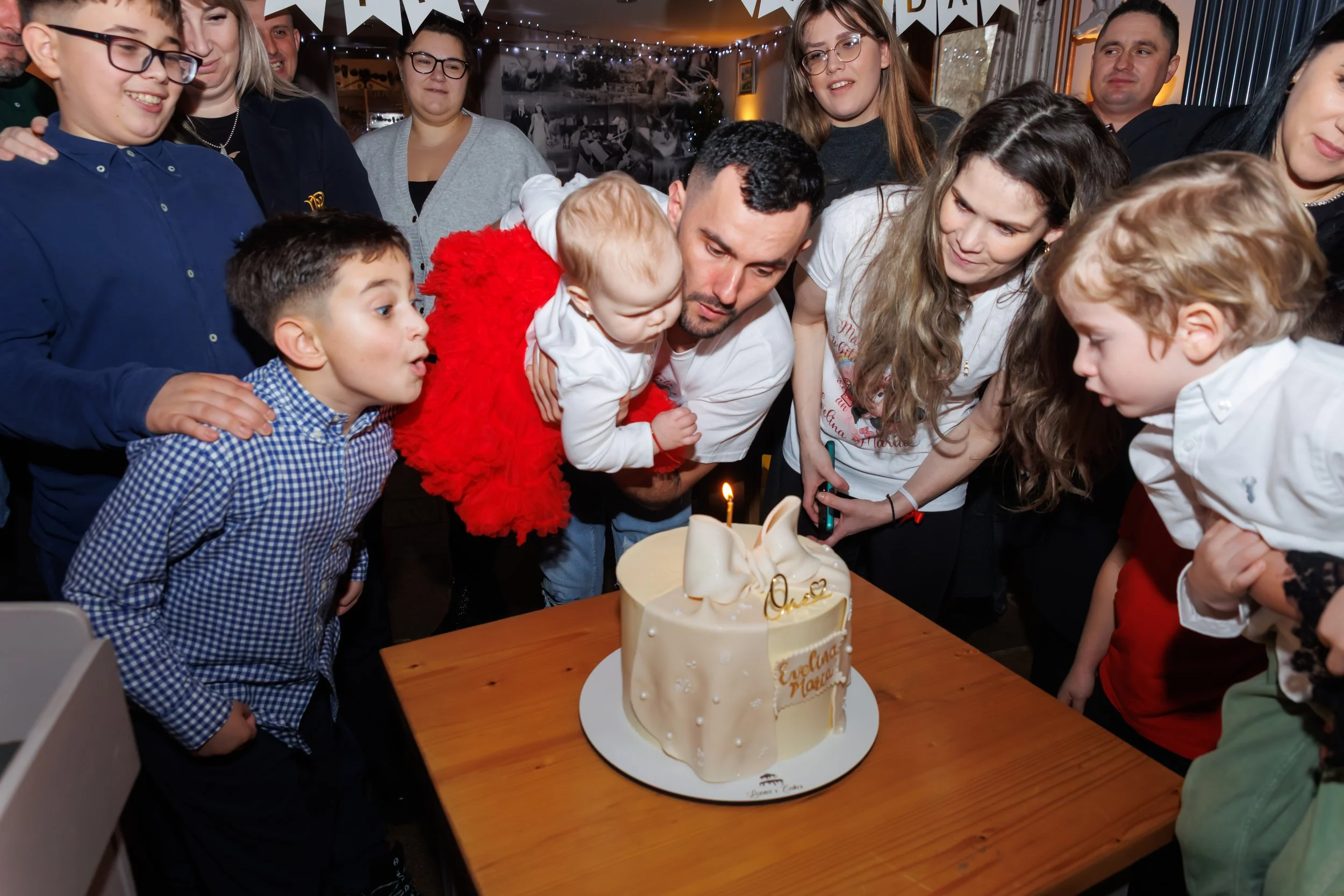 A group of children and adults gathered around a table, celebrating with a cake that has a single lit candle. The cake is decorated with white icing and a gold decal that reads 'Eternal Love.' The room has festive decorations including string lights and banners, indicating a birthday celebration.