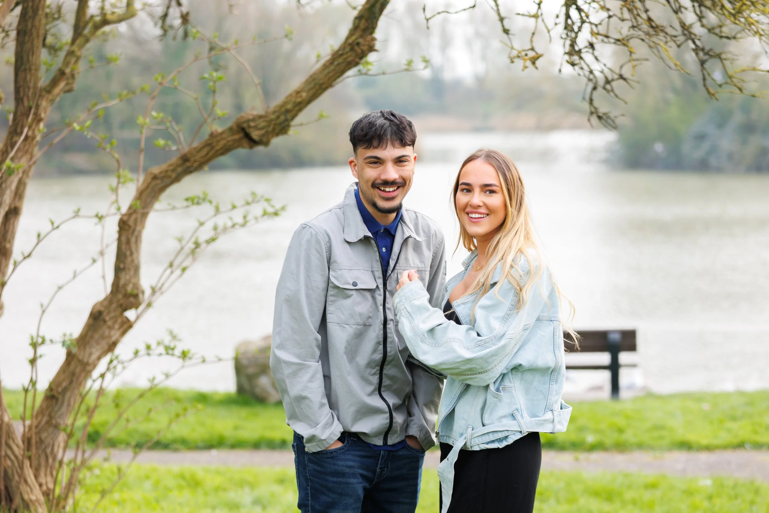 A happy young man and woman holding hands outdoors near a lake and trees, smiling at the camera.