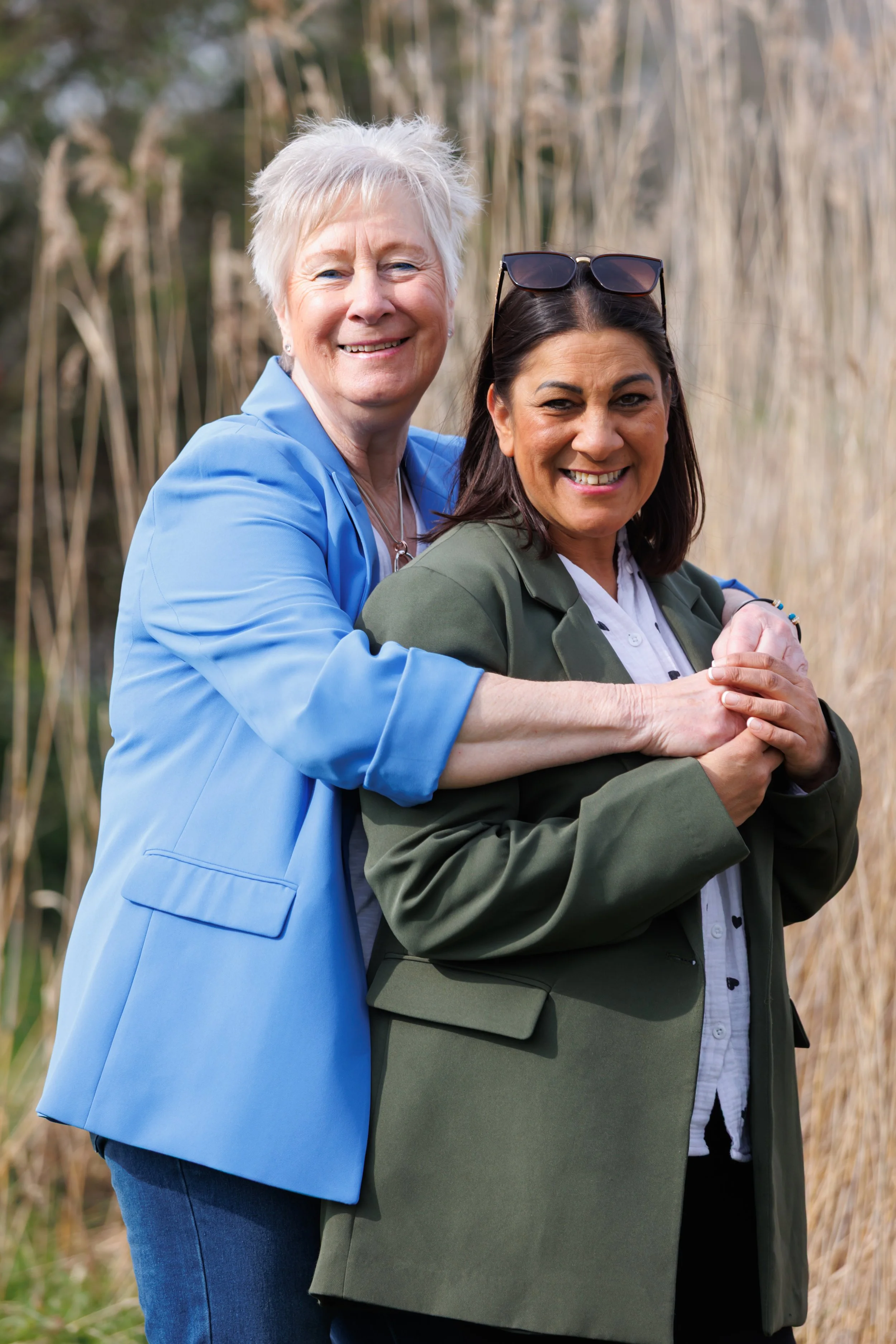 Two women smiling, standing outside among tall grass or reeds, embracing each other, with one woman wearing a blue jacket and the other a green blazer, and the woman in the blazer wearing sunglasses on her head.