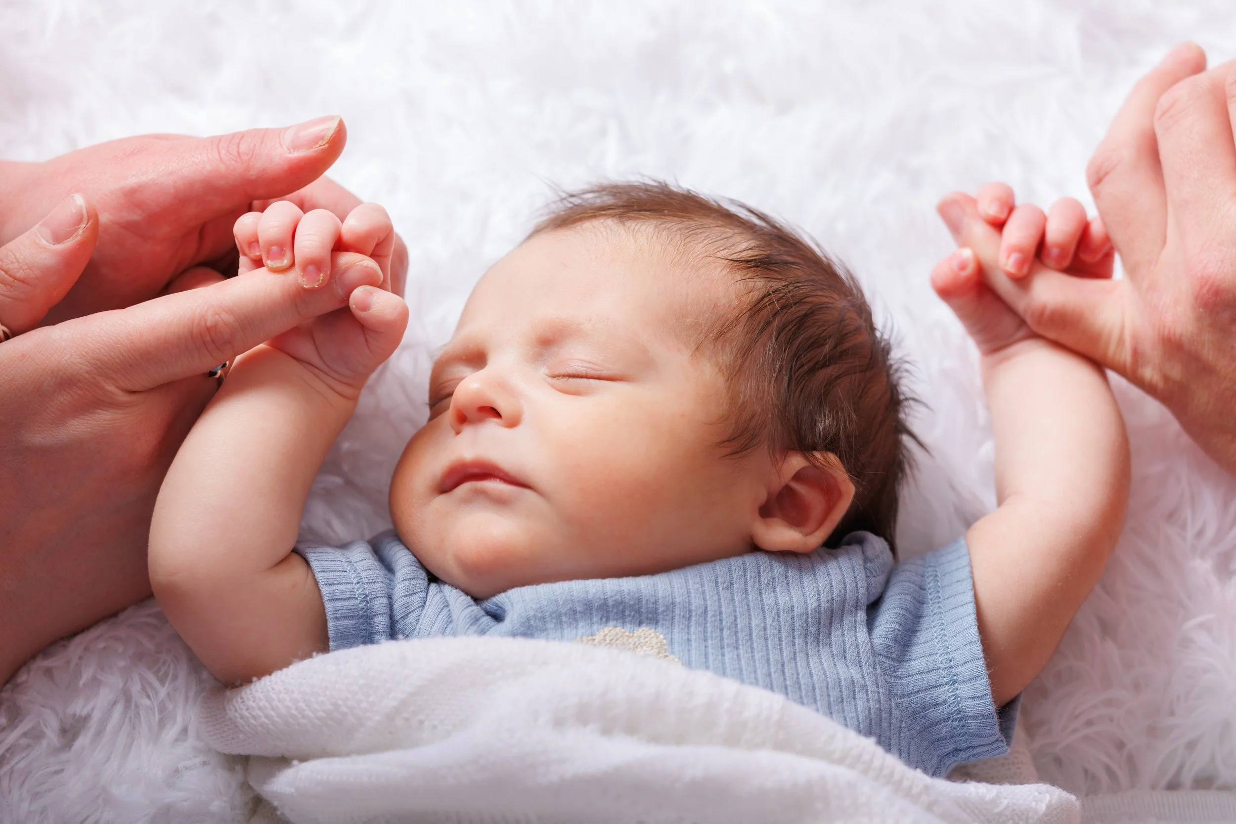 A baby lying on a soft, fluffy surface with adult hands gently holding the baby's hands.
