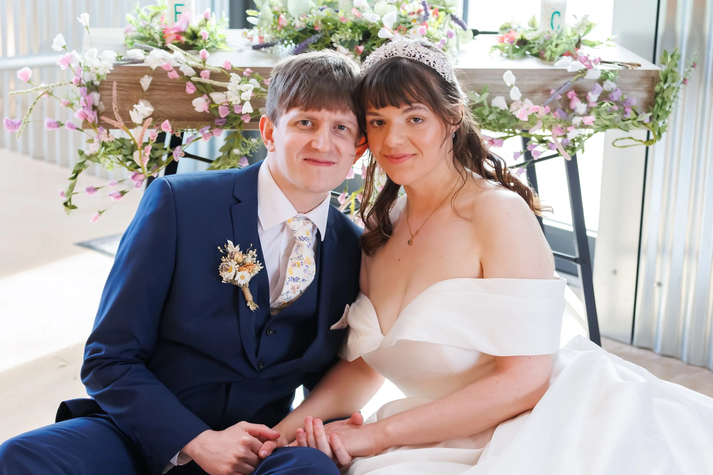 A newlywed couple sitting close together in front of a floral backdrop, smiling at the camera. The groom is wearing a dark blue suit with a floral tie and boutonniere, and the bride is in an off-the-shoulder white wedding gown with a tiara. They are 