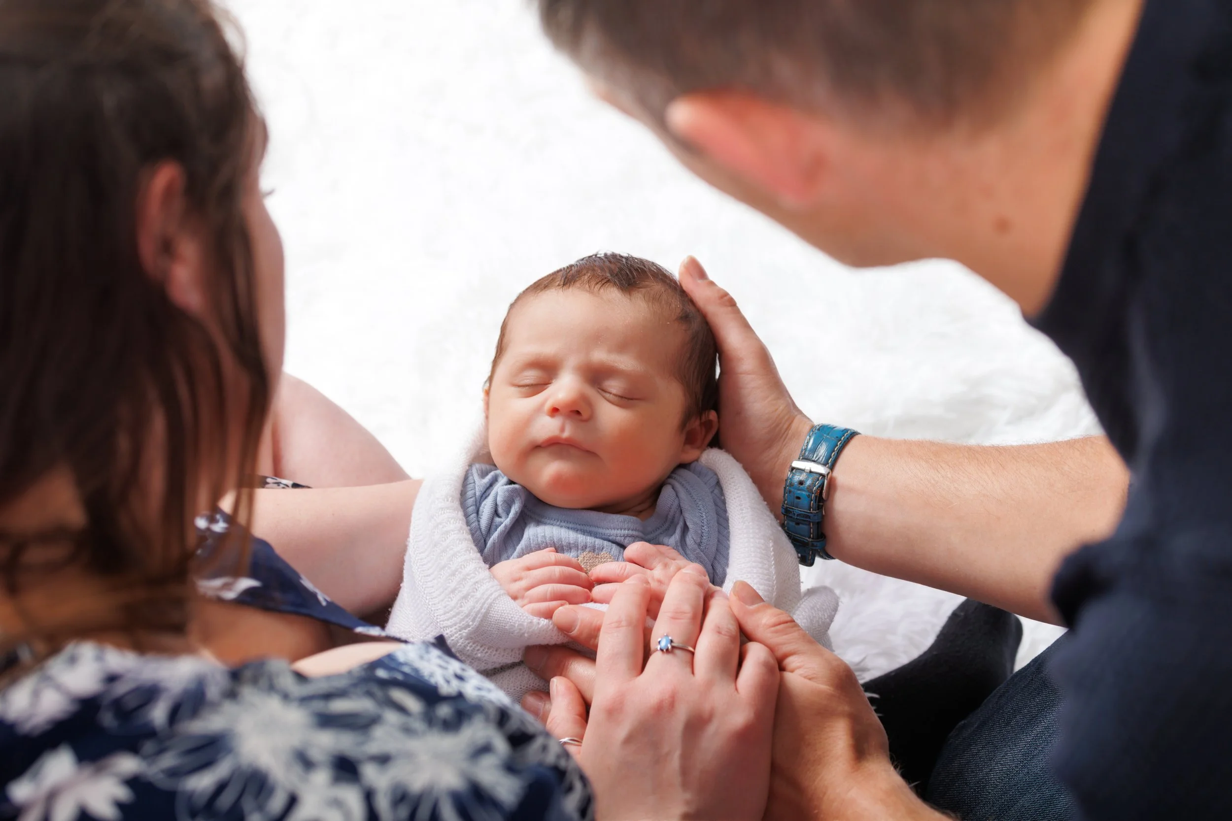 A family of three holding a newborn baby, with all faces close together, during a load moment. The baby is dressed in a white sweater and has eyes closed.