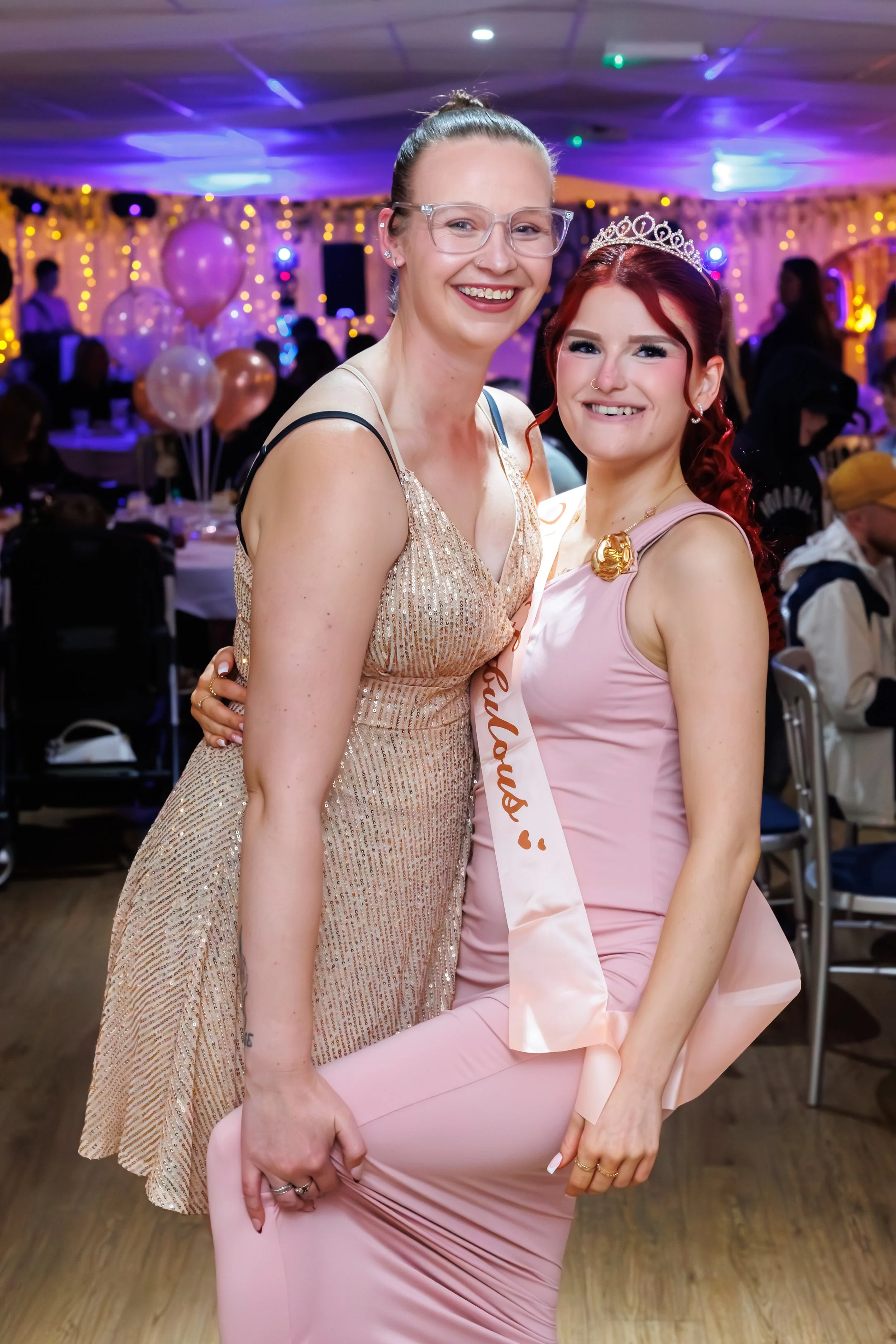 Two women smiling at a celebration, one wearing a pageant sash and a tiara, with indoor festive decorations, balloons, and other guests in the background.