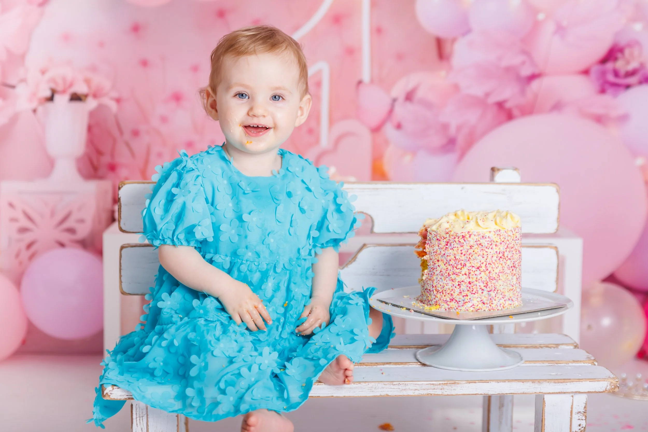 A smiling young girl with red hair and blue eyes, wearing a blue dress with flower patterns, sitting in front of a pink birthday cake with colorful sprinkles on a cake stand, at a birthday party with pink balloons and decorations in the background.