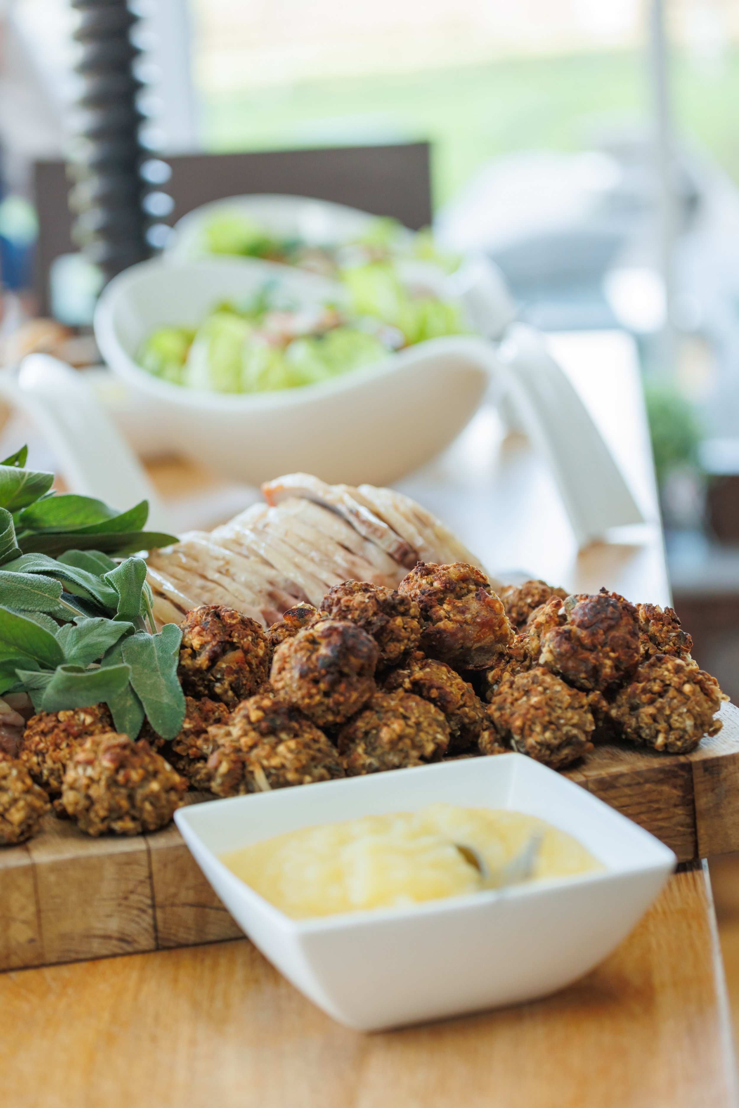 Close-up of a plate of cooked meatballs, fresh herbs, and slices of cooked chicken, with dipping sauce in a white bowl in the foreground and a bowl of green grapes in the background on a wooden surface.