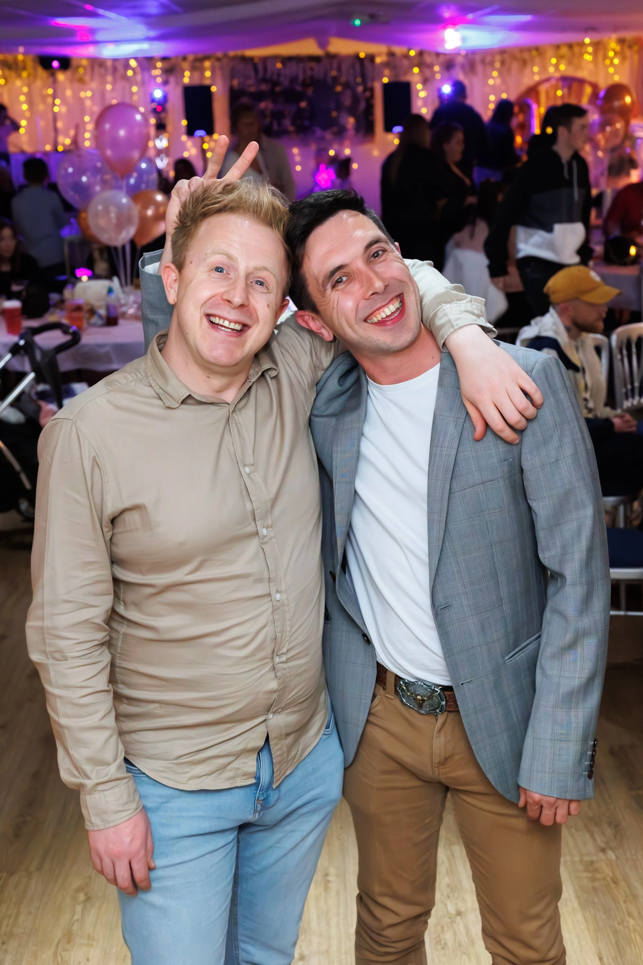 Two men smiling, one making a peace sign behind the other's head, at a lively indoor celebration with balloons, fairy lights, and other guests in the background.