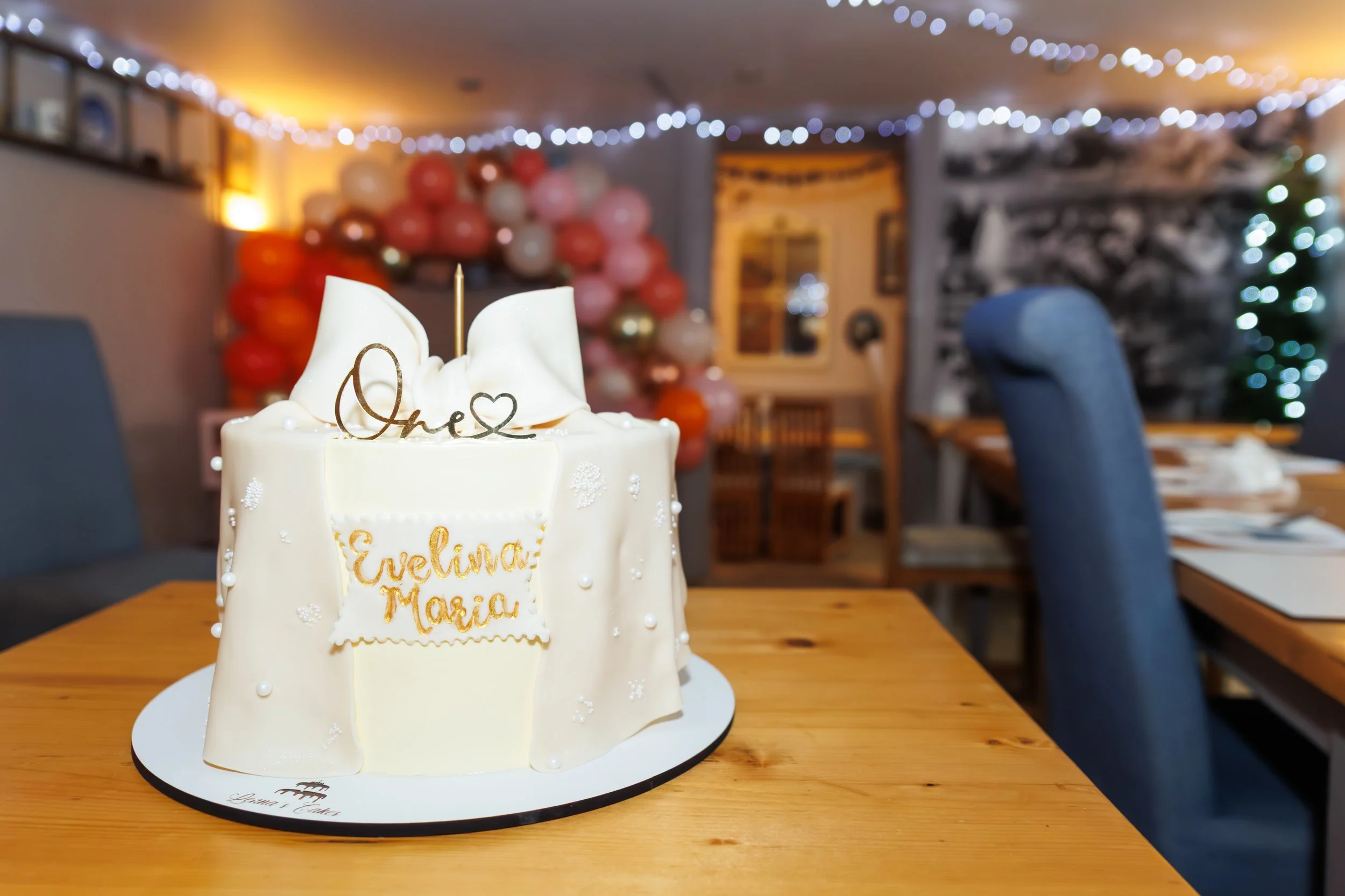 A white birthday cake with decorative gold writing that says 'Evelina Maria,' and a topper with the word 'One' and a heart symbol, placed on a wooden table in a decorated room with balloons and string lights.