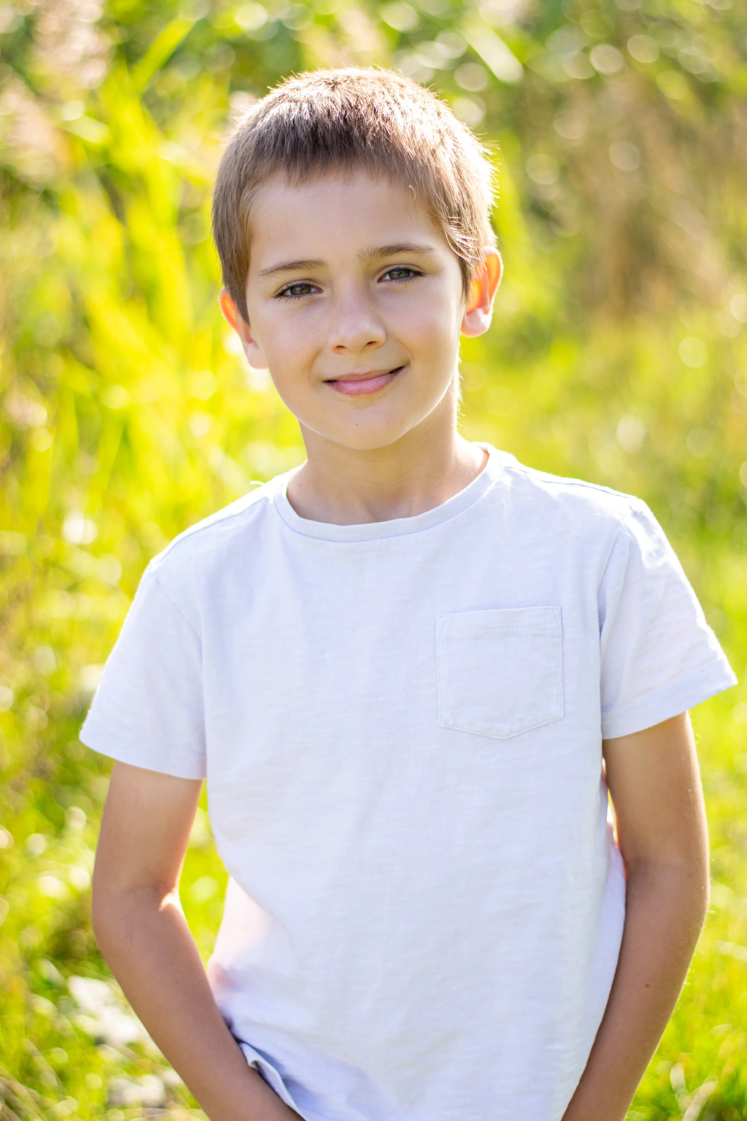 A young boy with short brown hair standing outdoors in a sunny, green, grassy area, wearing a white t-shirt.