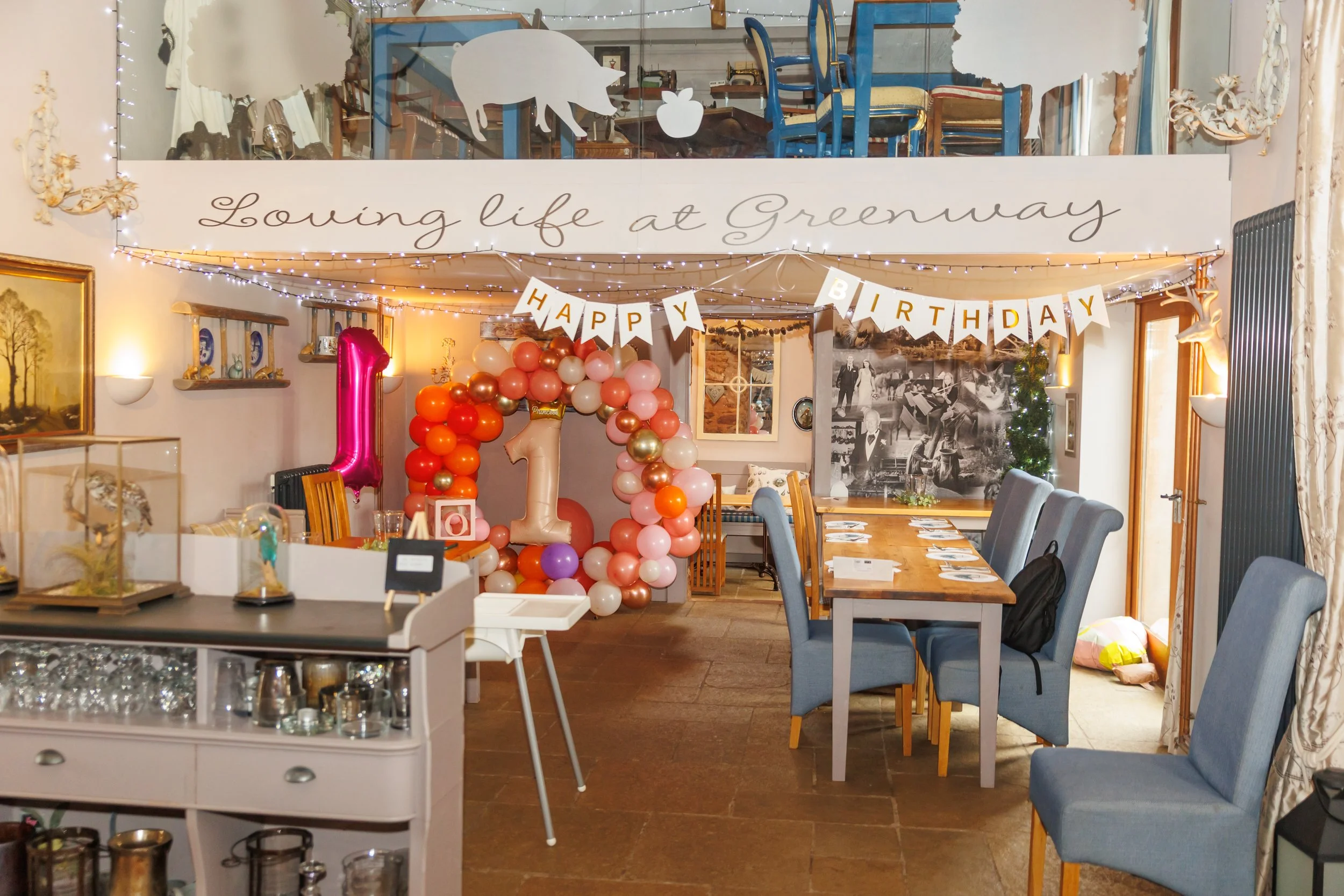 A celebration decorated with balloons, including a large pink number one balloon, and banners that read 'Happy Birthday'. The room has a sign that says 'Loving life at Greenway' and features various furniture, wall art, and fairy lights.