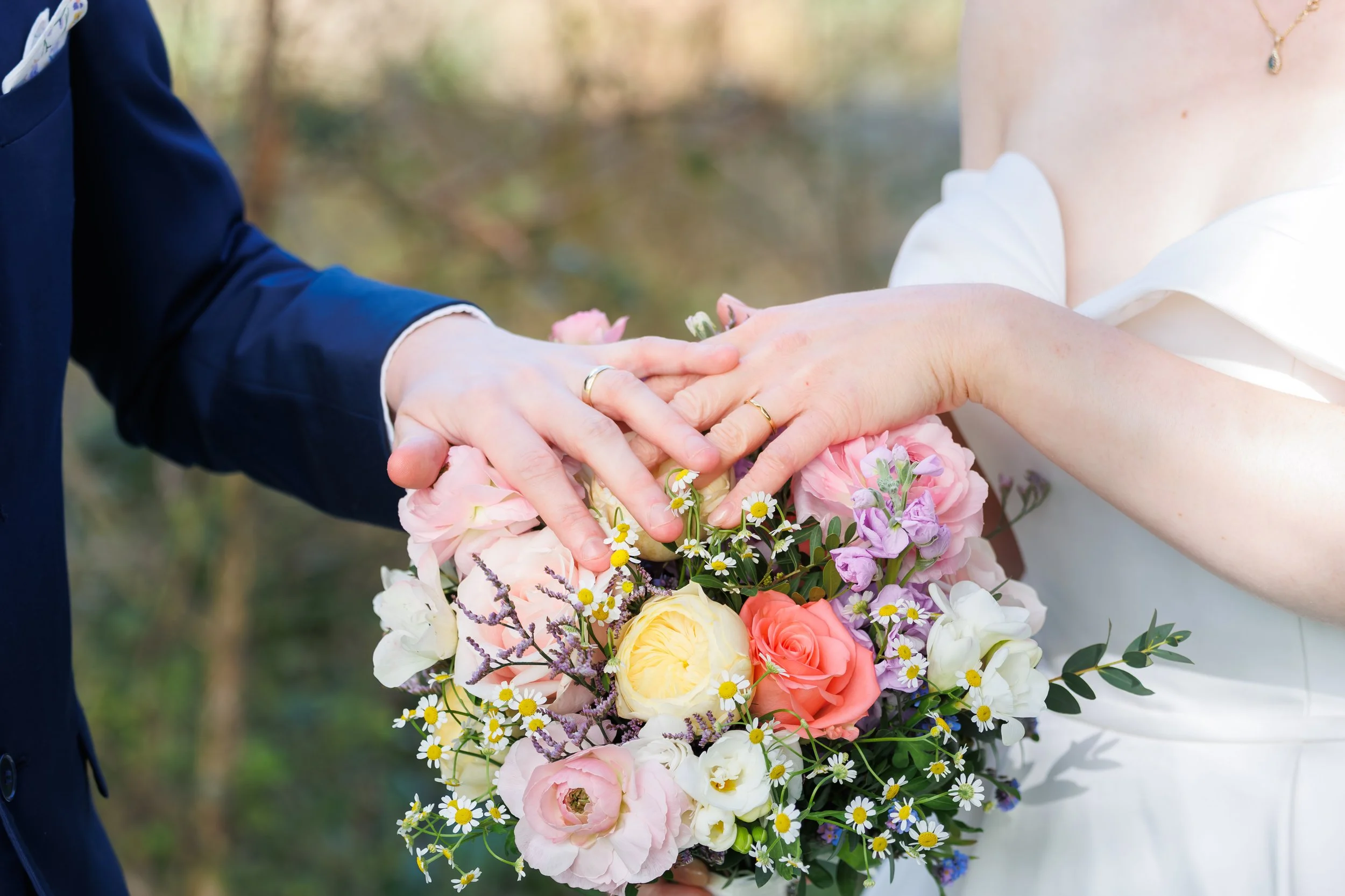 Close-up of a bride and groom holding hands during a wedding ceremony, with the bride holding a large, colorful bouquet of pink, yellow, white, purple, and small white daisies, and the groom wearing a navy blue suit.