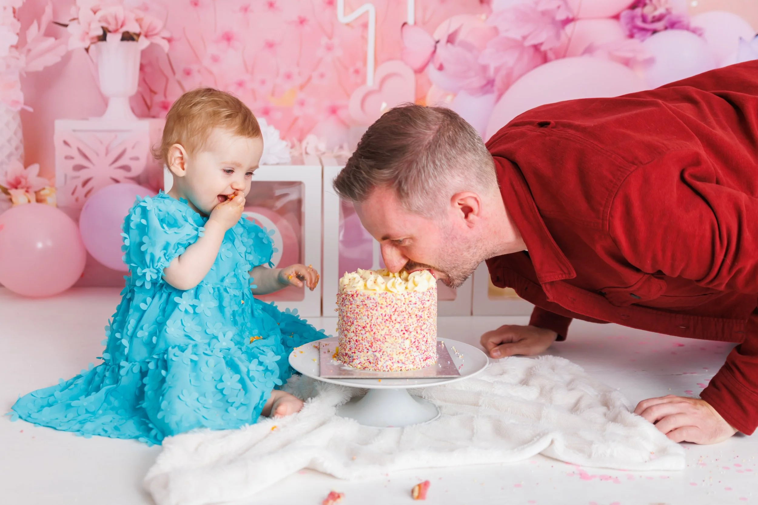 A young girl in a blue dress sitting on a fluffy white blanket, celebrating her birthday with a cake, while an adult man in a red shirt leans in to blow out the candles on the cake surrounded by pink balloons and decorations.