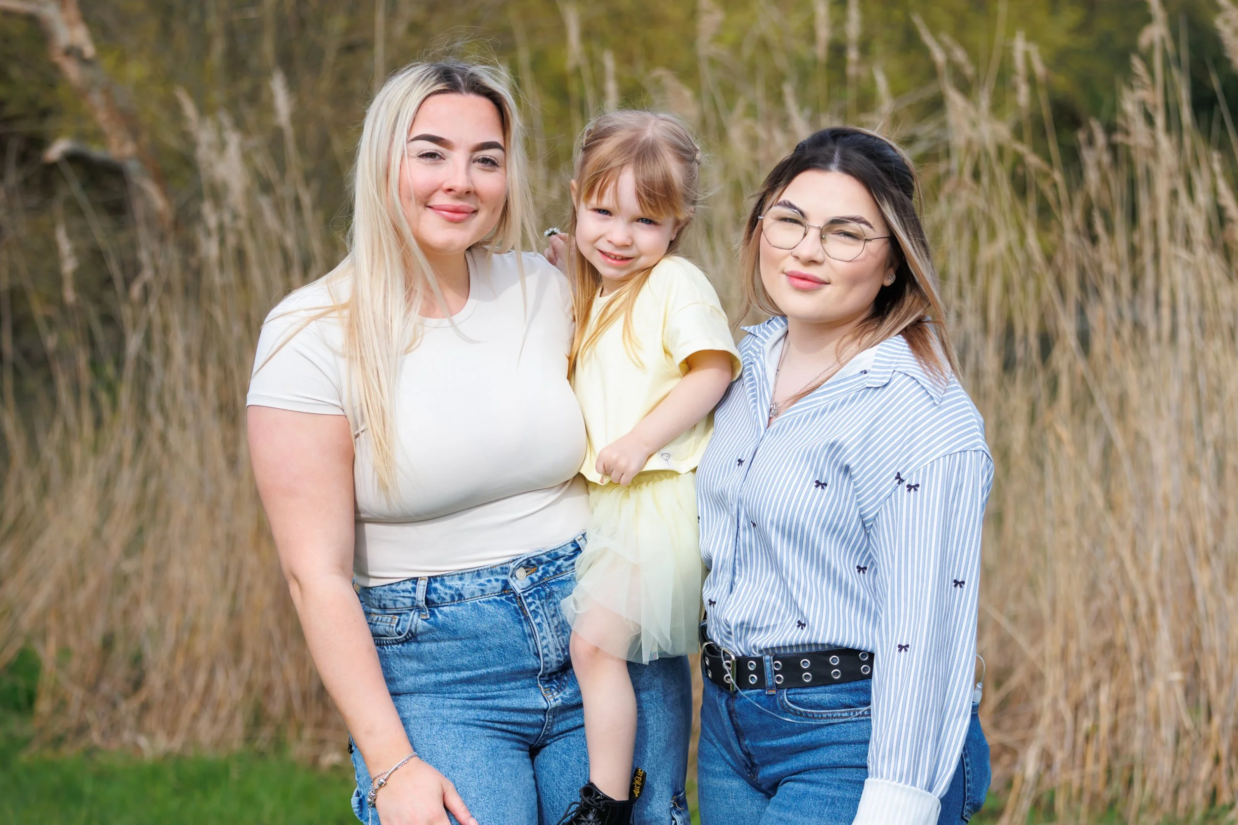 Three females and one young girl smiling outdoors in nature, surrounded by tall grasses and trees.
