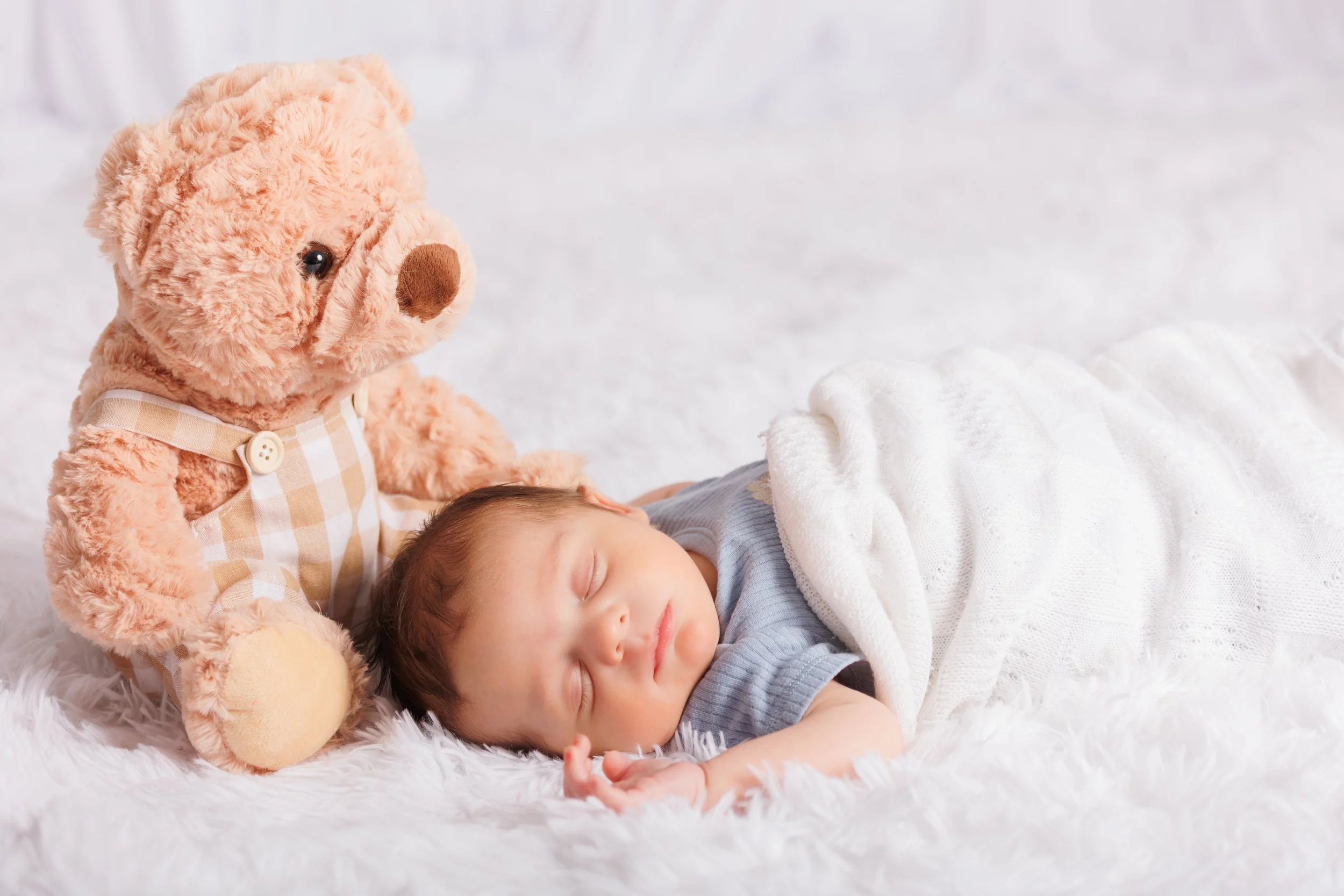 A young child with brown hair sleeping on a white blanket, with a plush teddy bear beside them on a fluffy white surface.