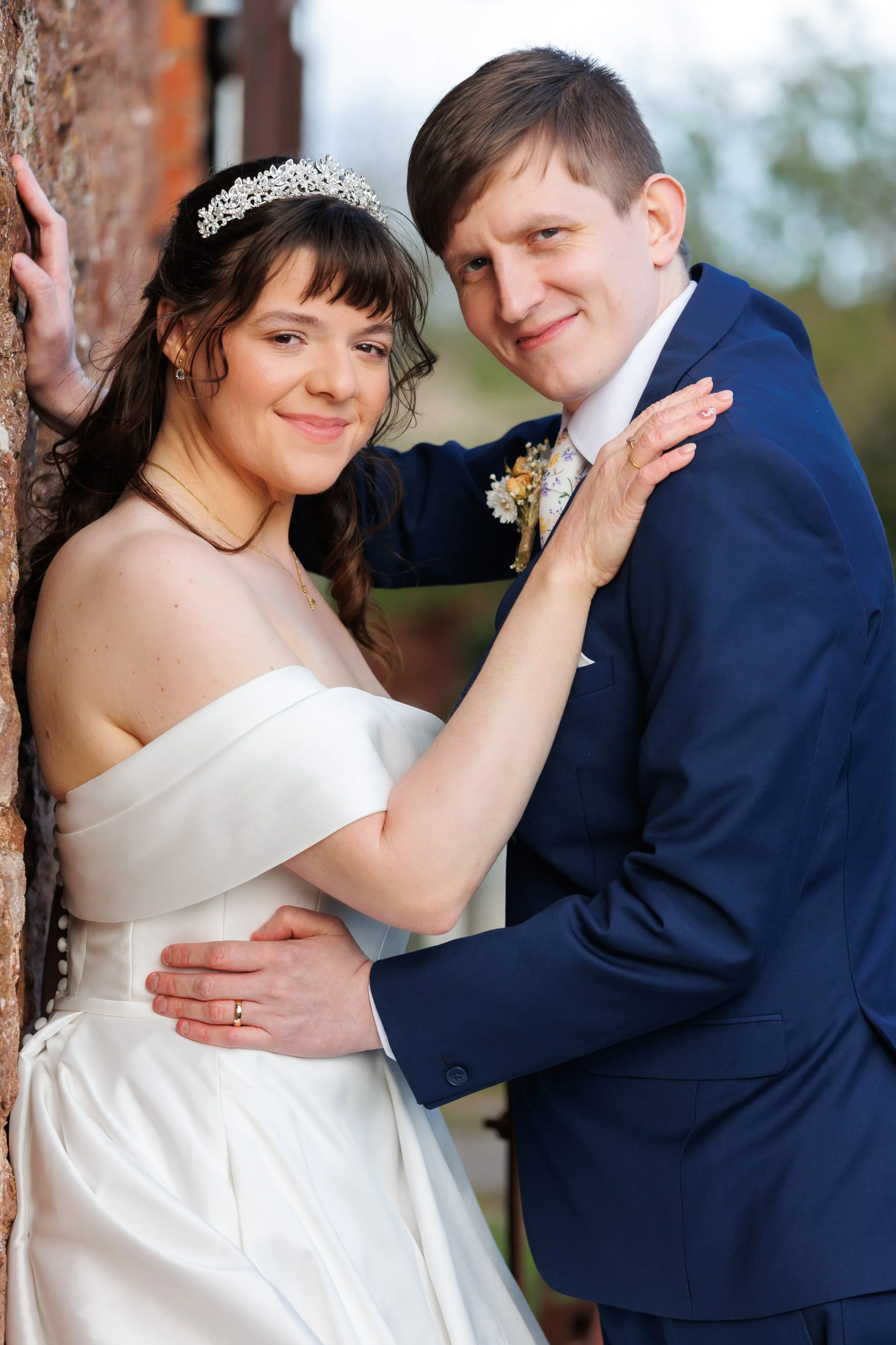 A newlywed couple on their wedding day, smiling and posing outdoors. The bride is wearing a white off-the-shoulder dress and a tiara, while the groom is dressed in a navy blue suit with a boutonnière. They are standing close with their hands gently touching.