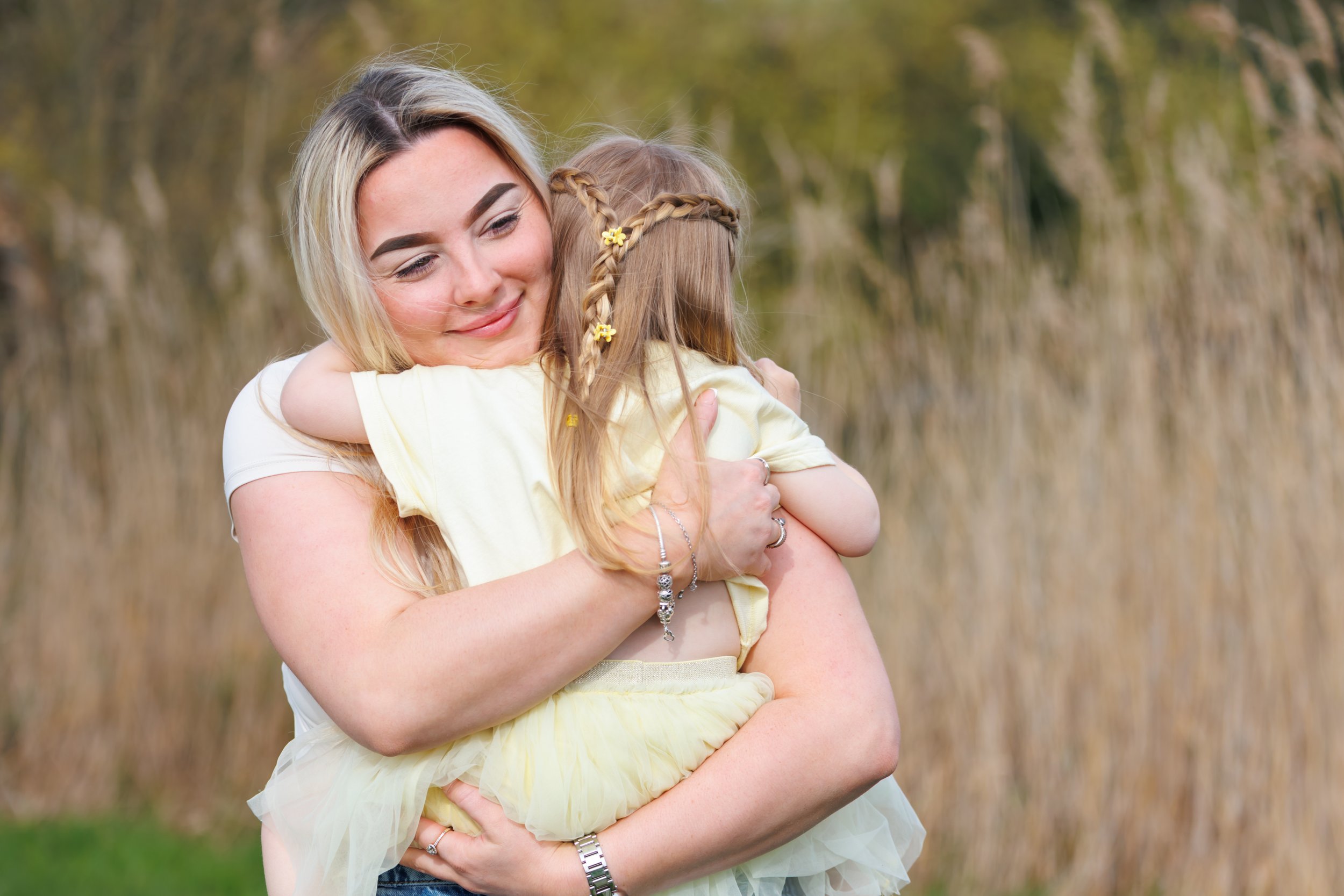 A woman with blonde hair holding a young girl with braided hair, outdoor setting with blurry trees in the background.