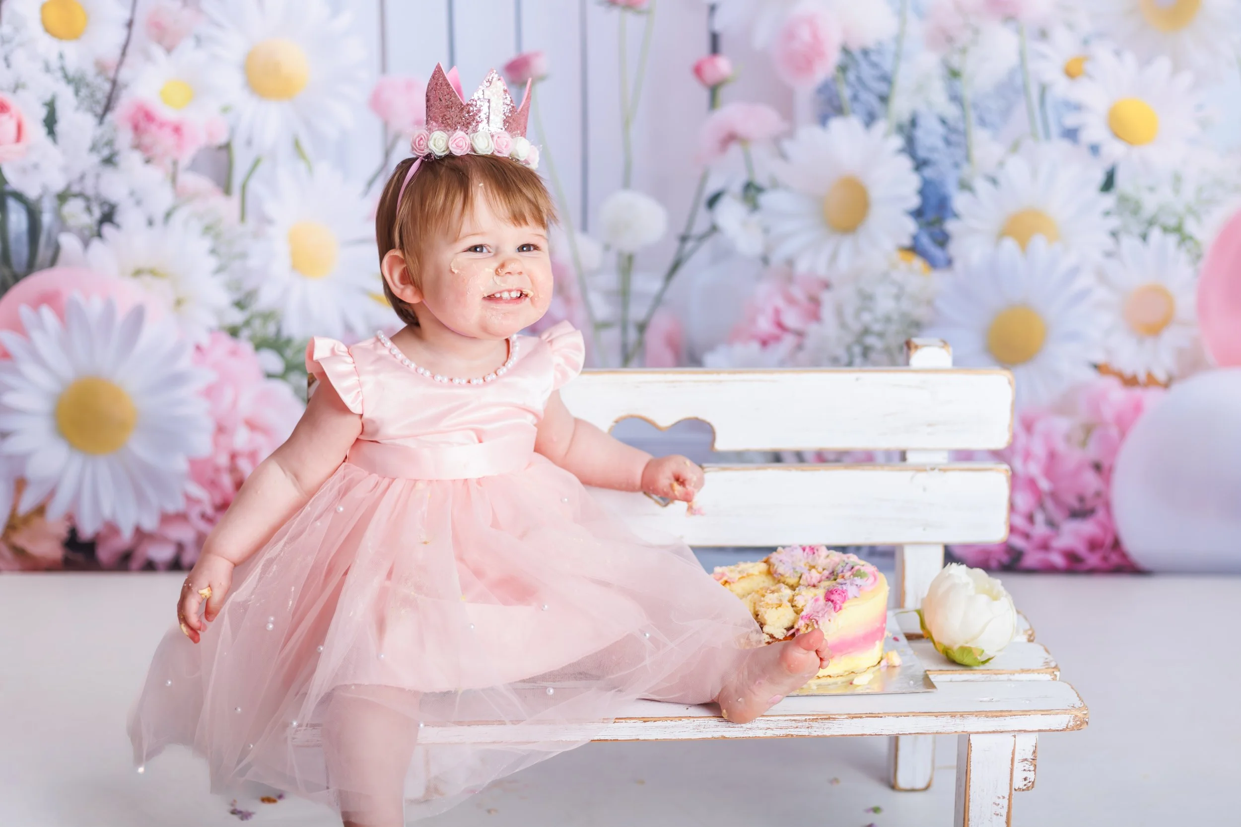 A small girl with a crown on her head, wearing a pink dress, is sitting at a cake table with cakes and strawberries. The background is decorated with large white daisies and pink flowers.