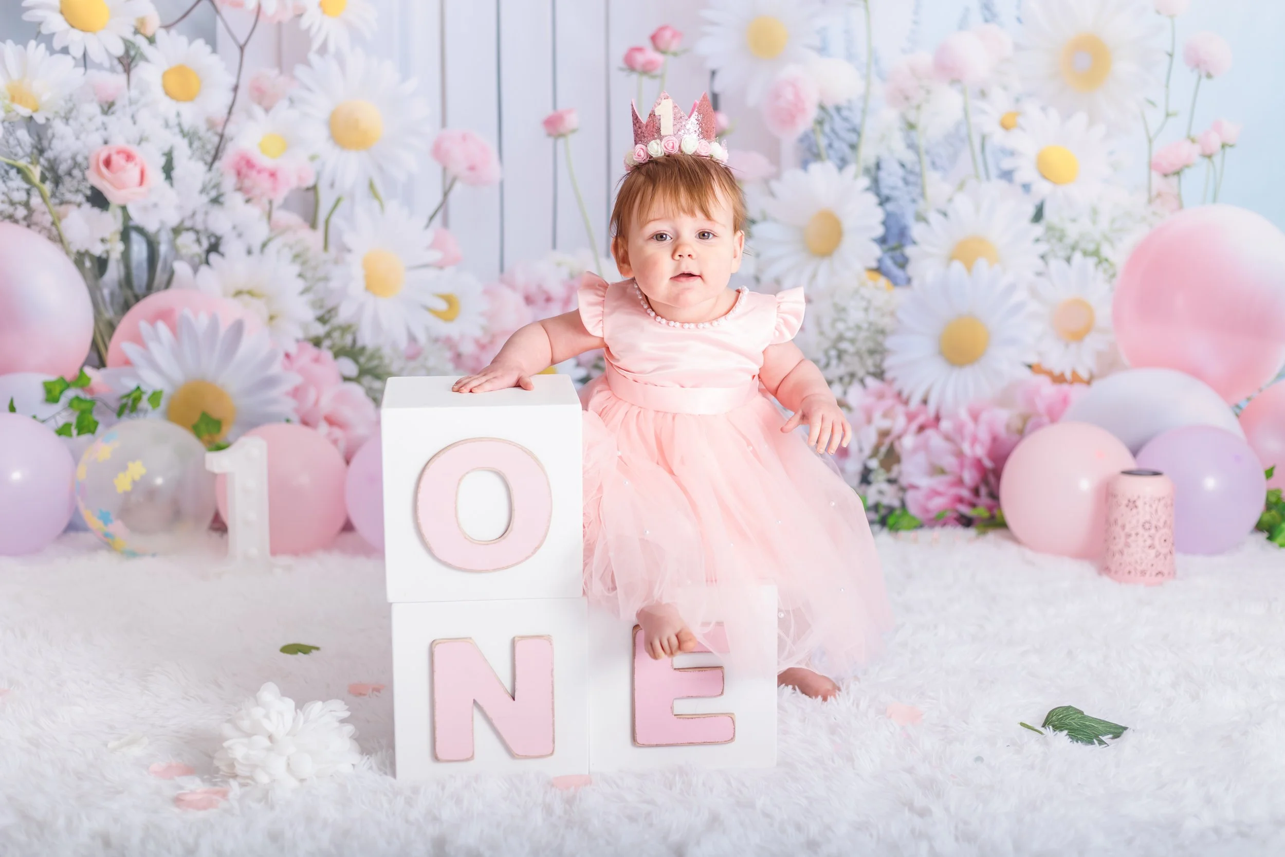 A young girl wearing a pink dress and a birthday crown, sitting in front of a floral backdrop with balloons and flowers, celebrating her first birthday with decorative blocks spelling 'ONE'.