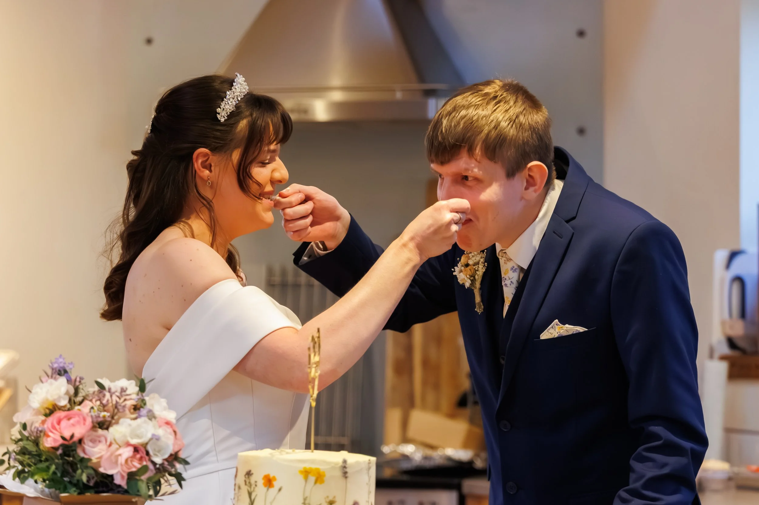 A bride and groom at their wedding reception. The groom is pretending to pick his nose while the bride is smiling and laughing. The bride is holding a bouquet of pink, white, and purple flowers. There is a white wedding cake with floral decorations i