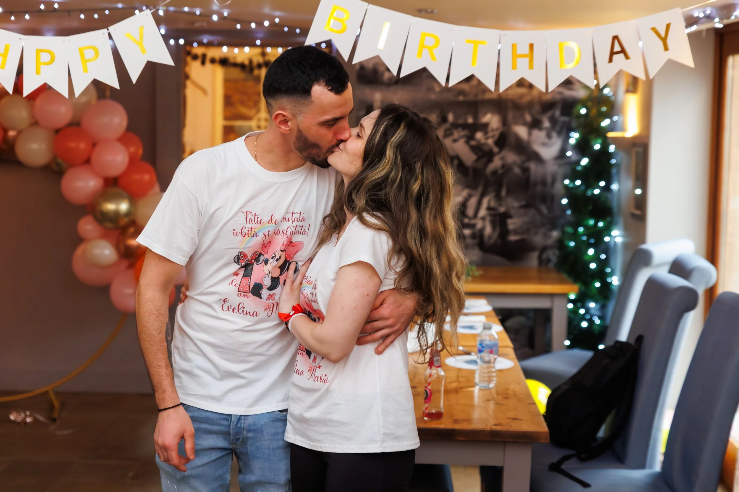 A couple shares a kiss at a birthday celebration with 'Happy Birthday' banners, balloons, and decorations in the background.