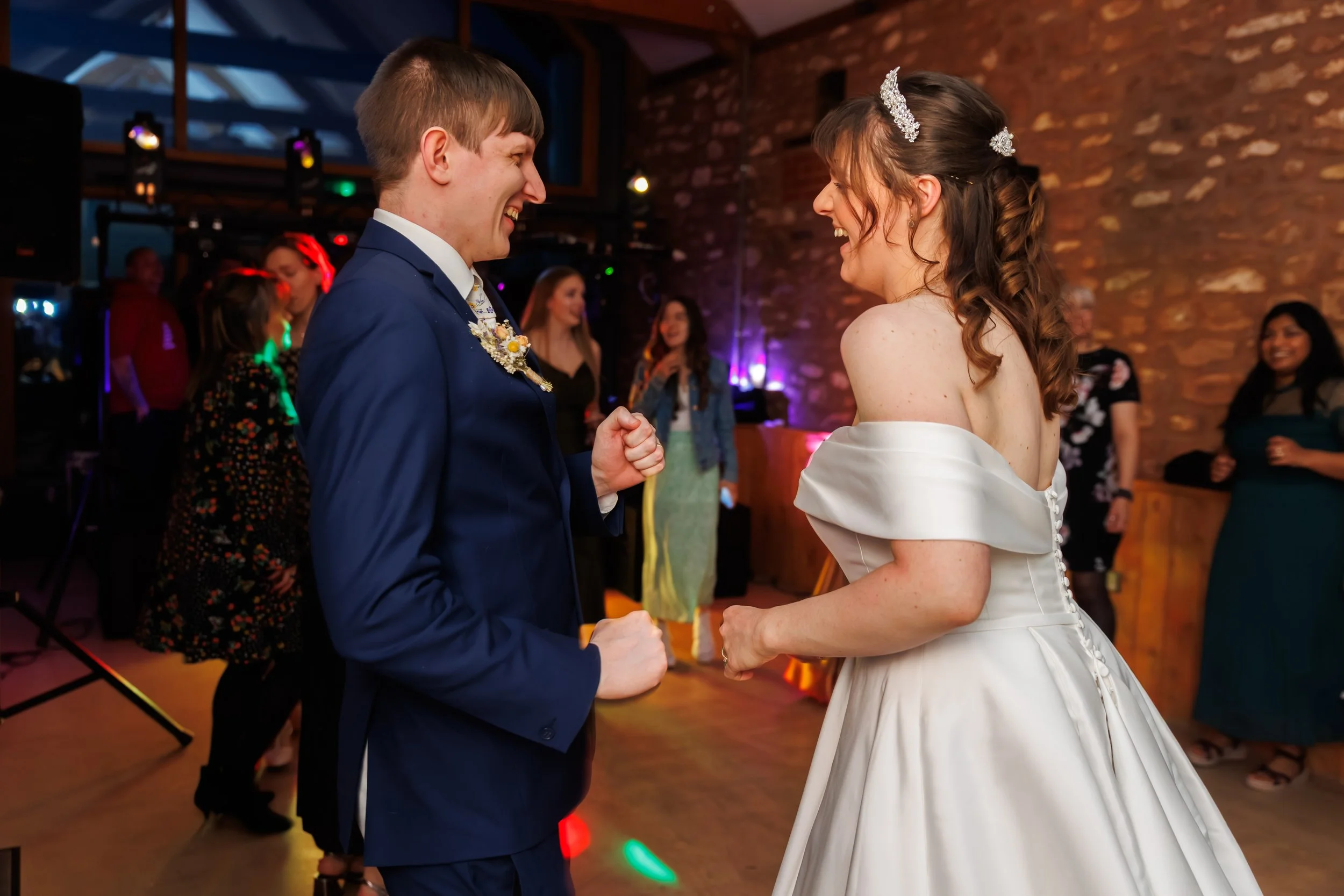 A man in a blue suit and a woman in a wedding dress are dancing and smiling at each other at a wedding reception, with other guests in the background.