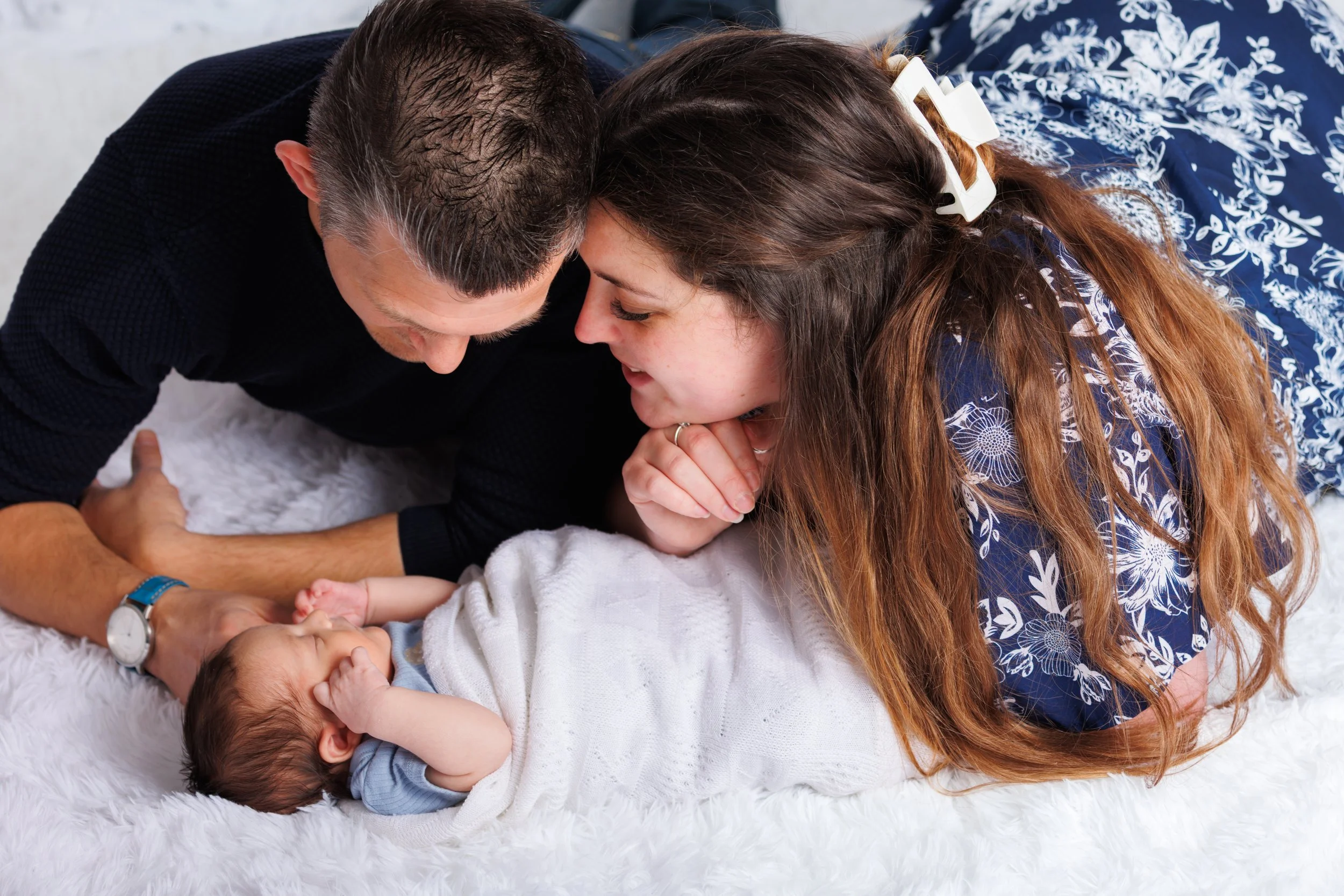 A family on the floor with a newborn baby. The father and mother are touching the baby's hands and foreheads, creating a tender moment, on a white furry rug.