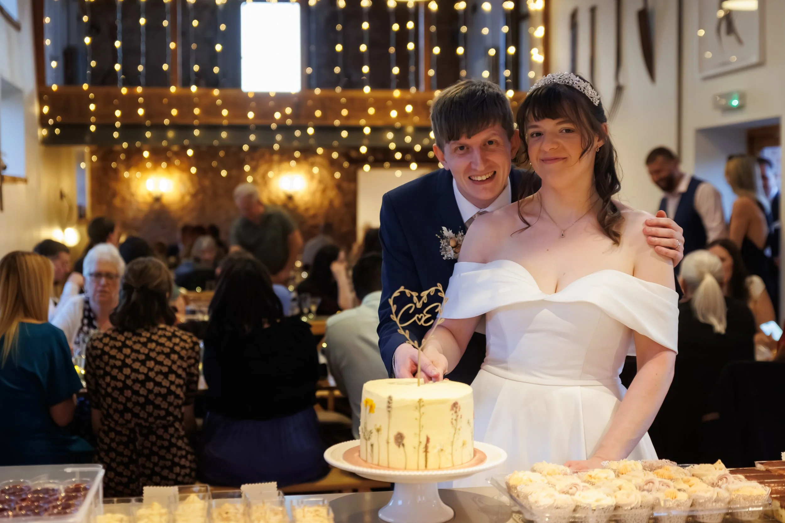 A newlywed couple at their wedding reception, with the groom wearing a suit and the bride in a white wedding dress and tiara, smiling at the camera, standing behind a layered cake on a table with other desserts, in a decorated hall with warm lighting