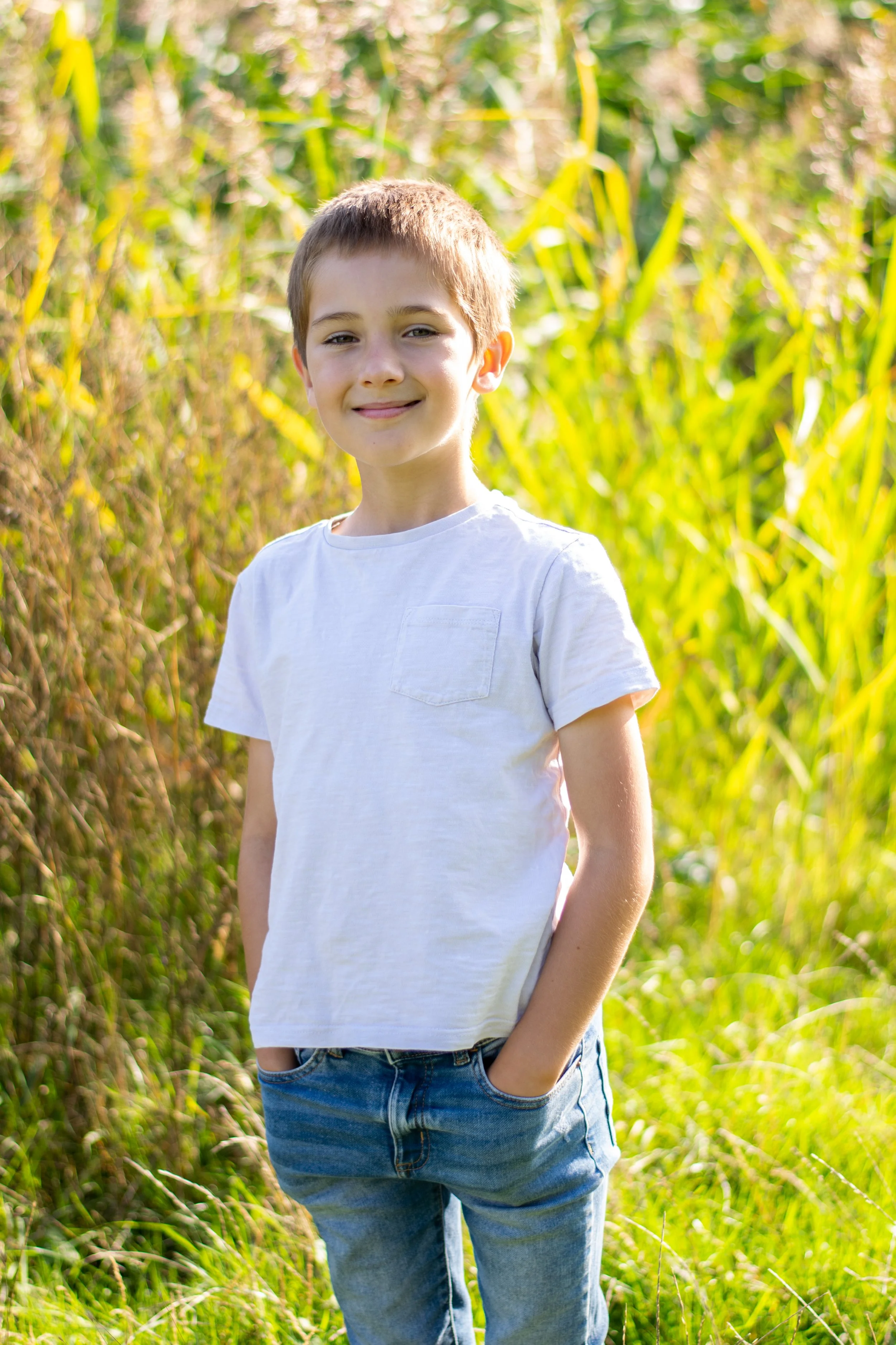 A smiling young boy with hands in pockets standing outdoors in a grassy field with tall green plants.