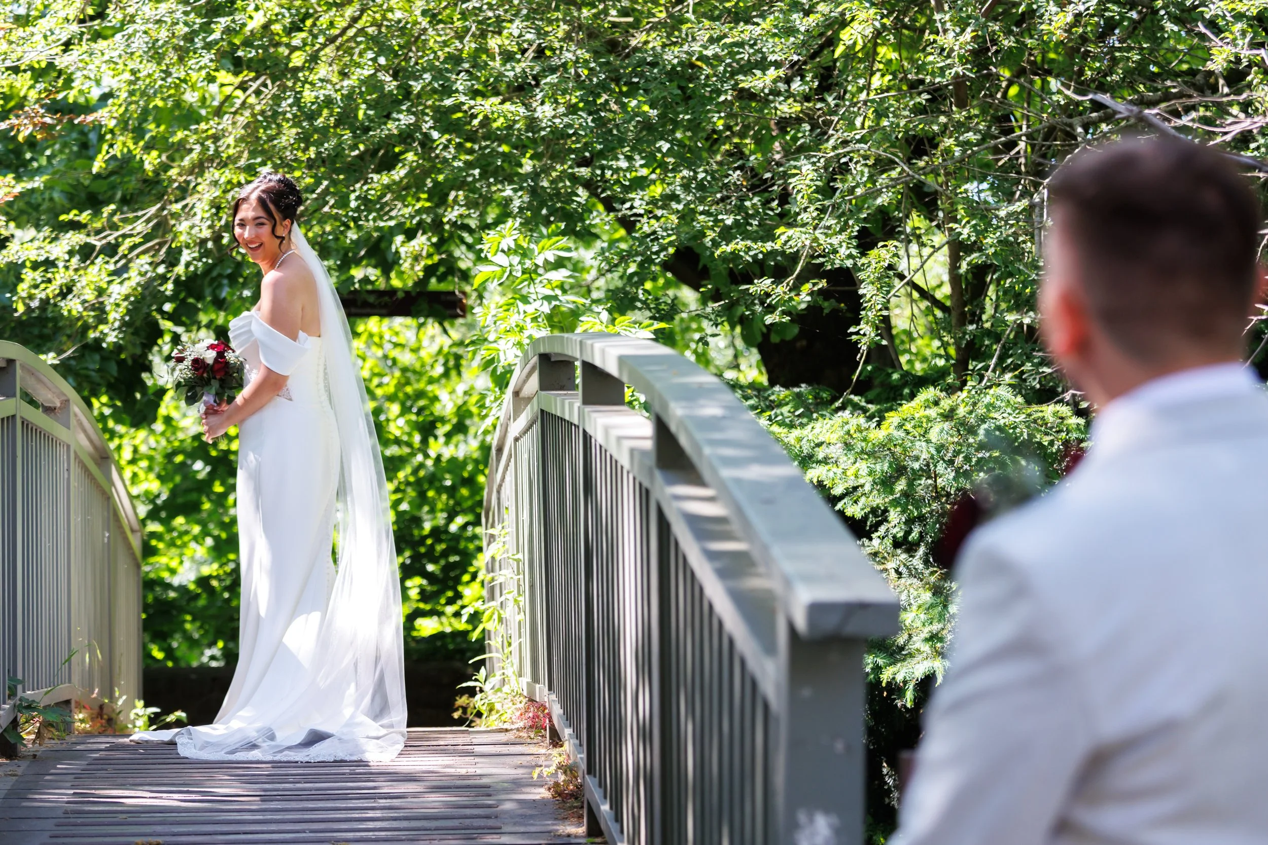 A bride in a white dress holding a bouquet, smiling, standing on a wooden bridge surrounded by lush green trees, with a man in the foreground looking at her.