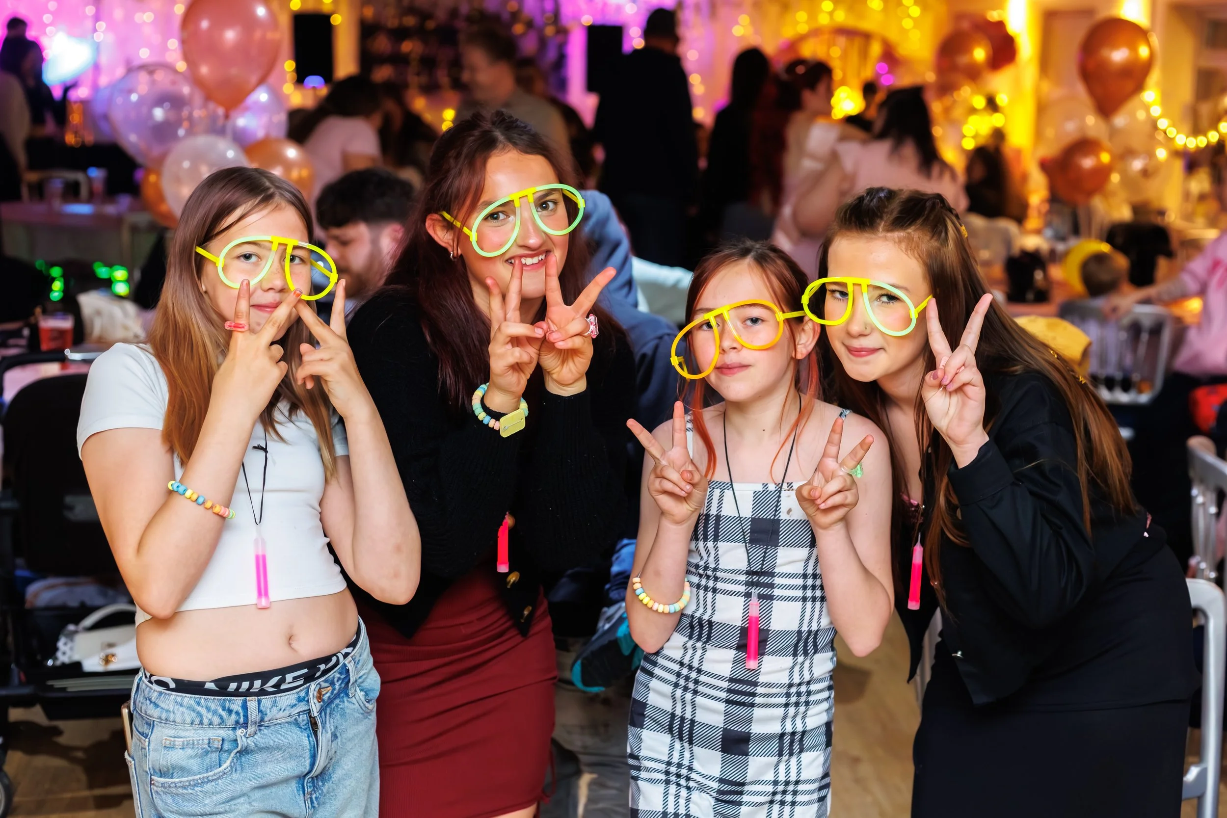 Four young girls wearing glow-in-the-dark glasses and making peace signs, posing at a lively party with balloons and colorful lights in the background.