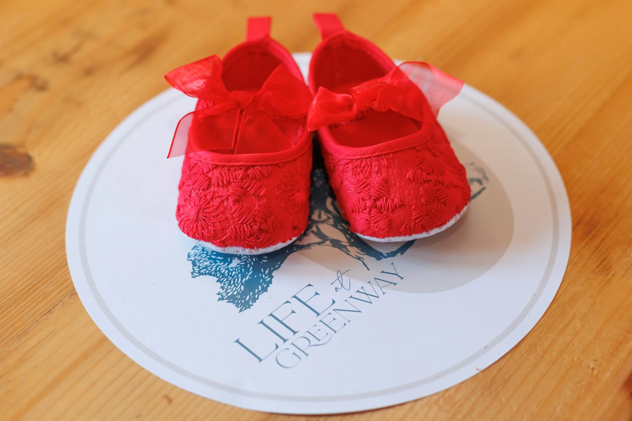 A pair of red baby shoes with decorative bows placed on a round white paper with blue writing and a tree illustration, on a wooden surface.