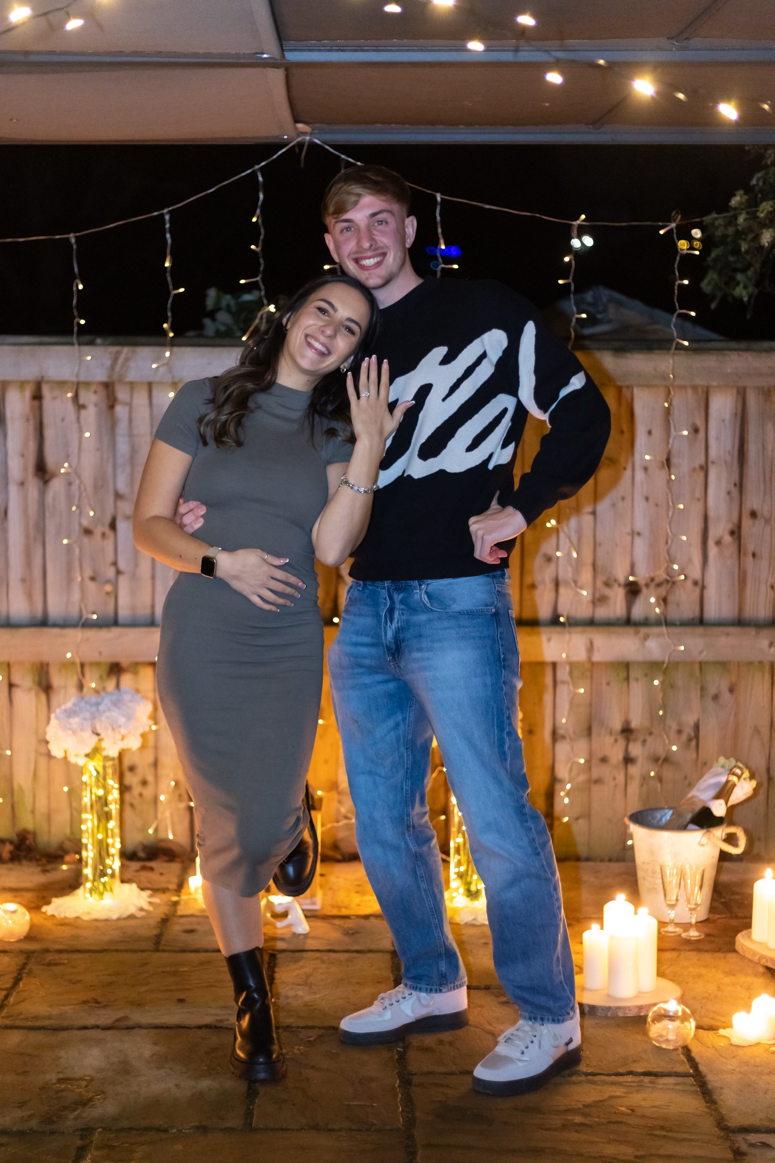 A couple celebrating with a wedding ring, surrounded by candles and string lights in an outdoor setting at night.
