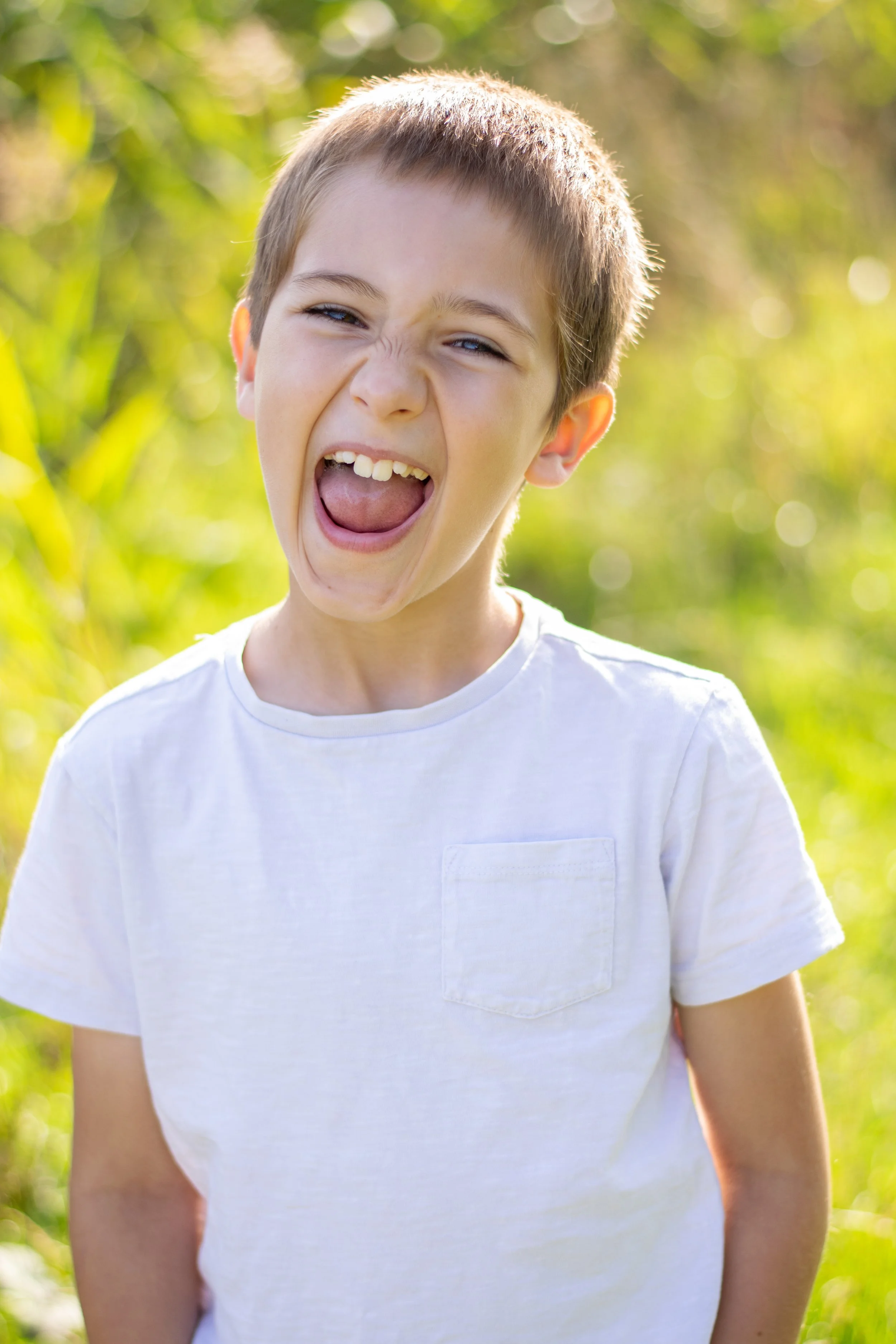 A young boy with light brown hair and blue eyes, wearing a white t-shirt, is outdoors, making a playful face with his mouth open wide, in front of a blurred background of green foliage.