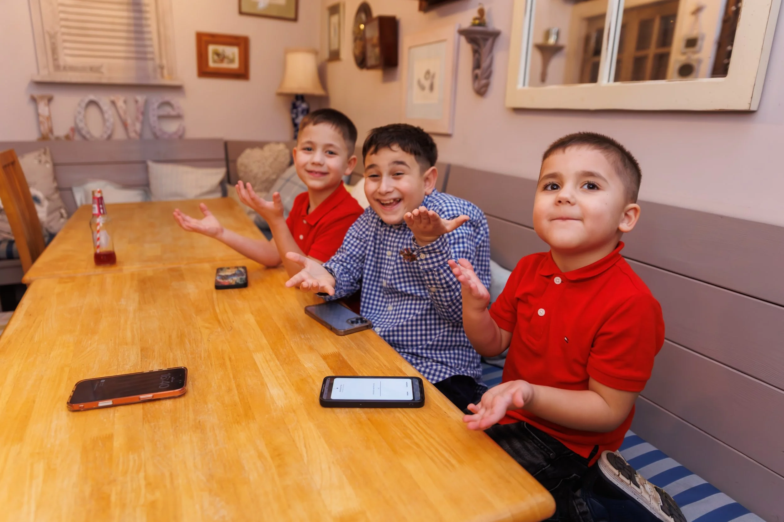 Three young boys sit at a wooden table with smartphones in front of them, smiling and gesturing with their hands. The background includes framed pictures, a lamp, and decorative items on the wall.