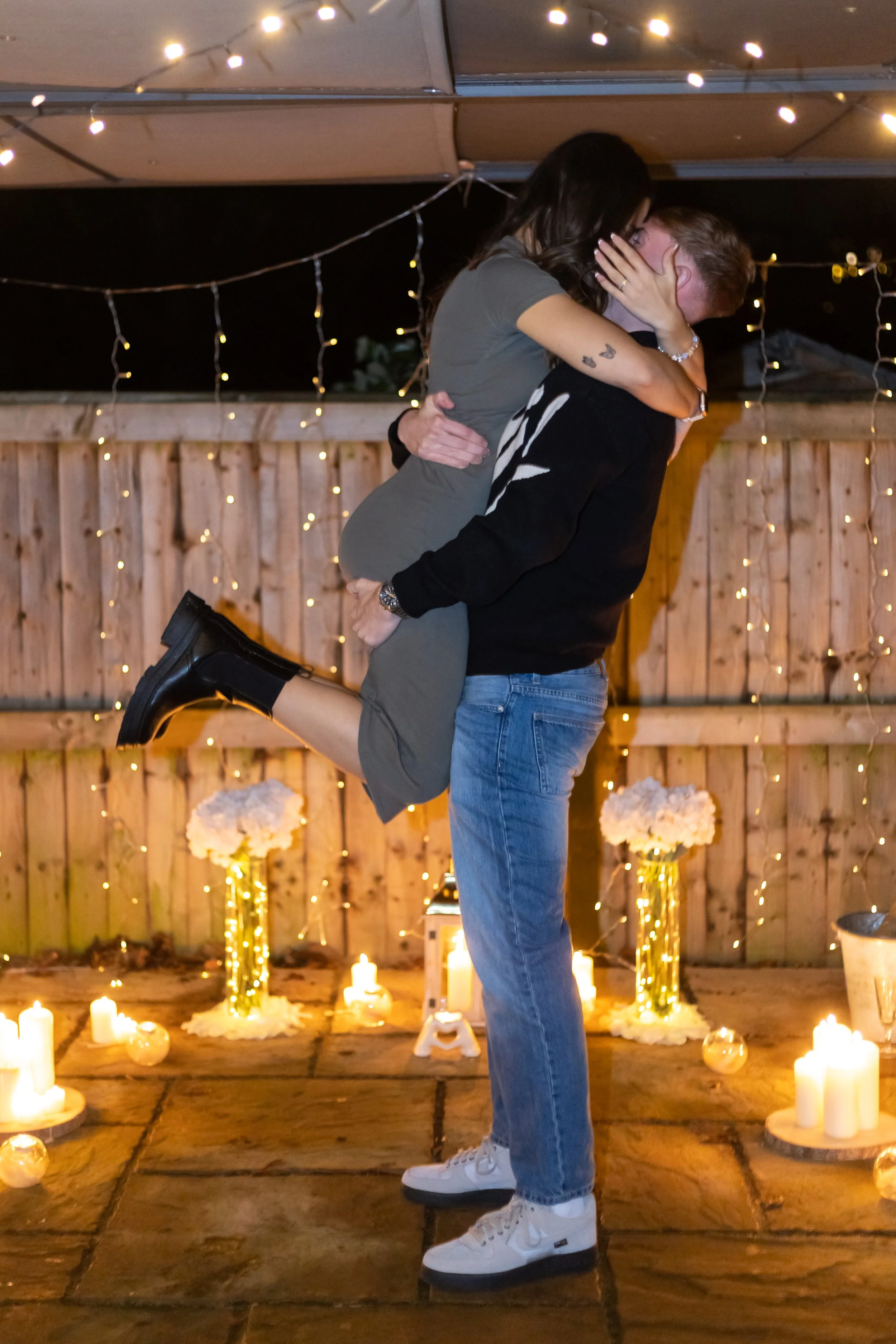A couple embraces in a romantic setting at night, with the man lifting the woman off the ground. The background is decorated with string lights, candles, and flowers on the ground, creating a warm and intimate atmosphere.