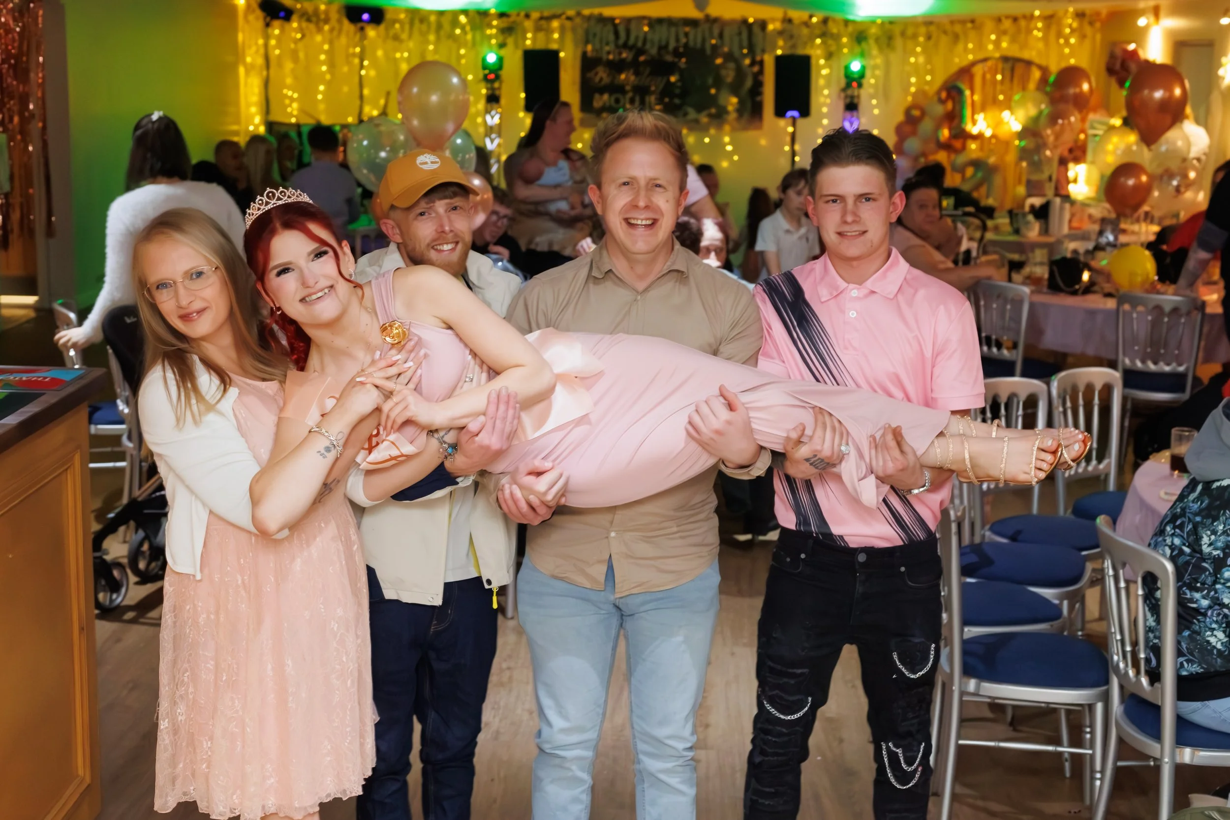 Group of five people at a celebration or party, with one person being held horizontally by the others. The background is decorated with balloons, fairy lights, and other colorful decorations, indicating a festive atmosphere.