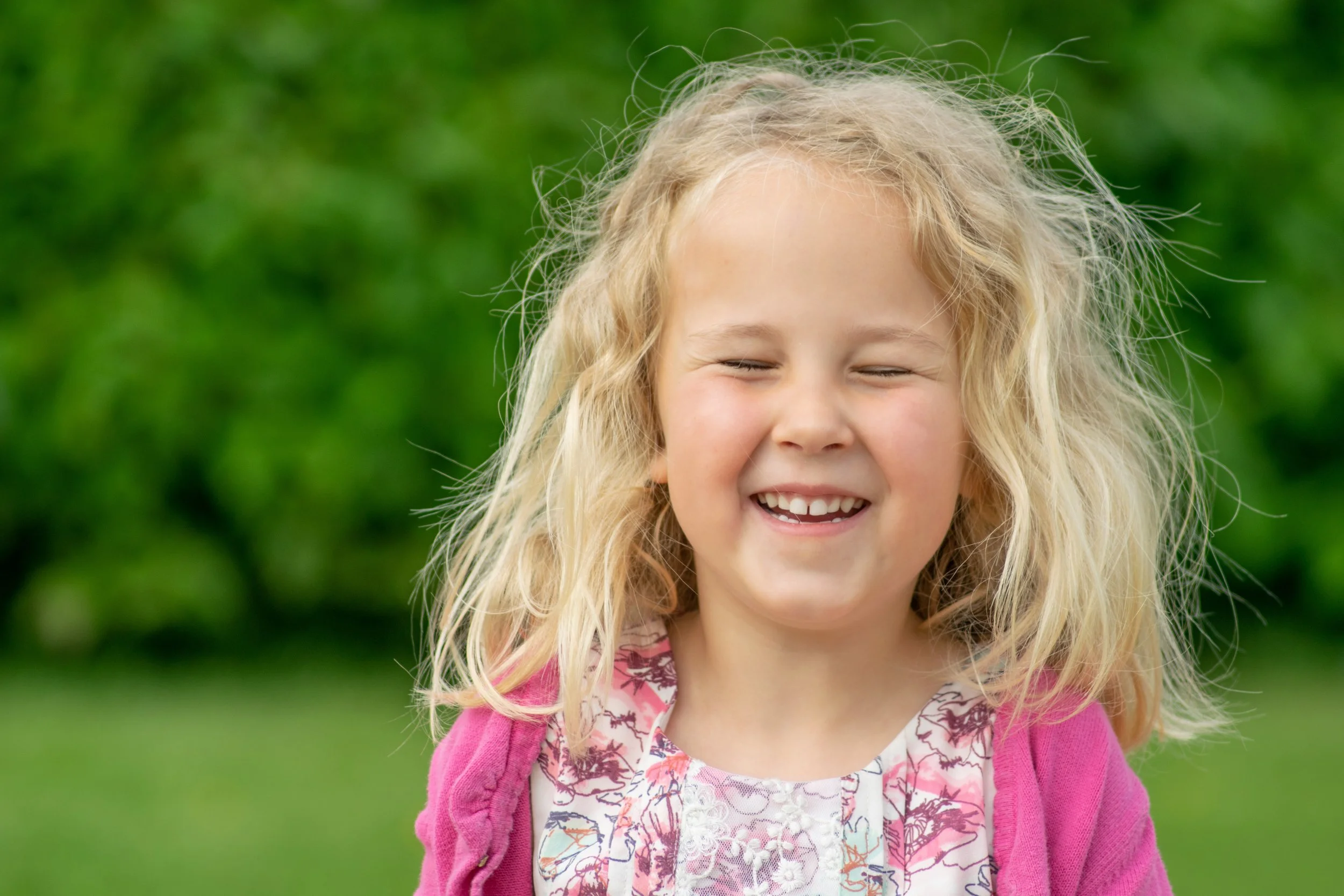 A young girl with blonde curly hair smiling with her eyes closed, wearing a pink cardigan and a floral blouse, standing outdoors with a blurred green background.
