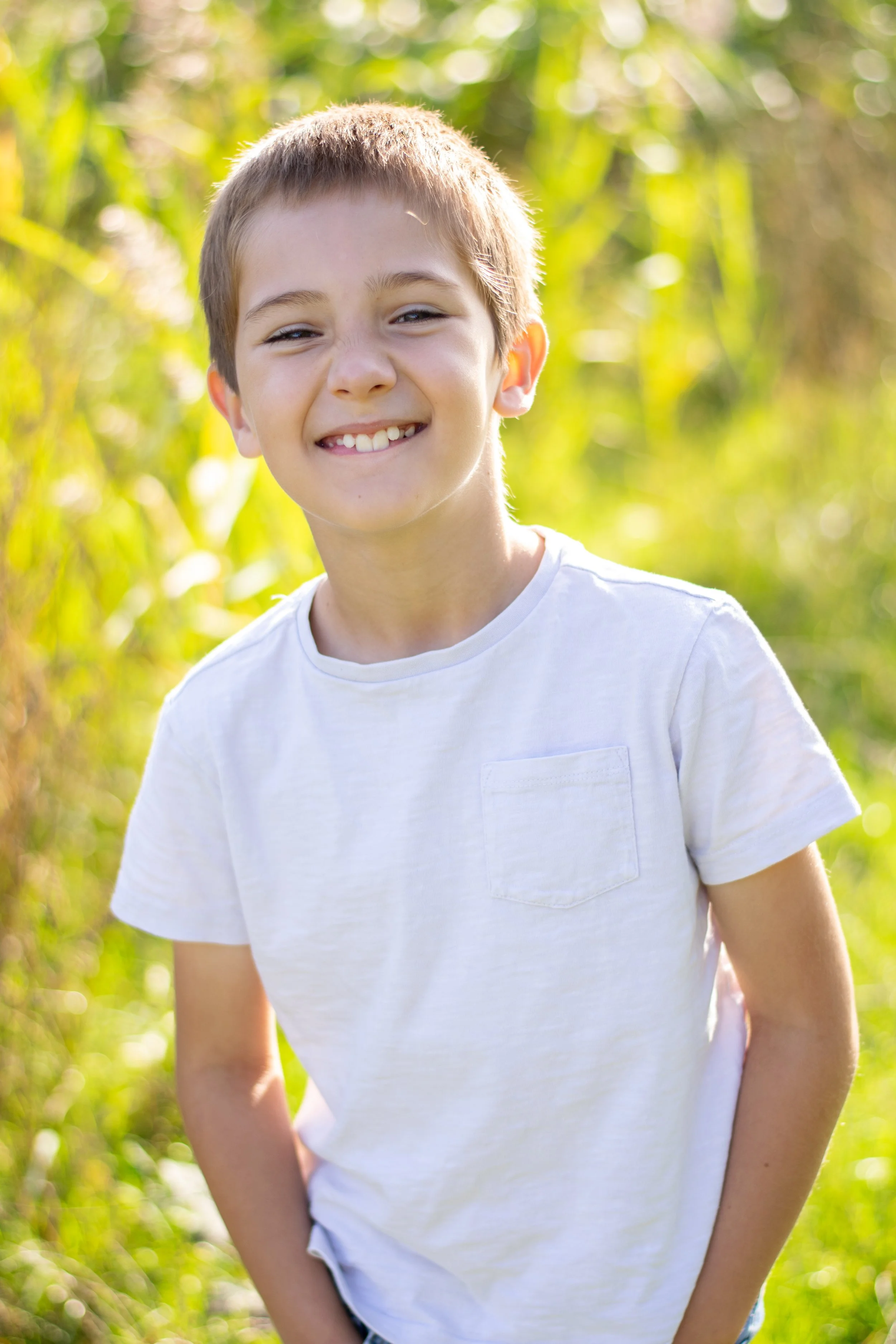 A smiling boy with short brown hair wearing a white T-shirt outdoors with green foliage in the background.