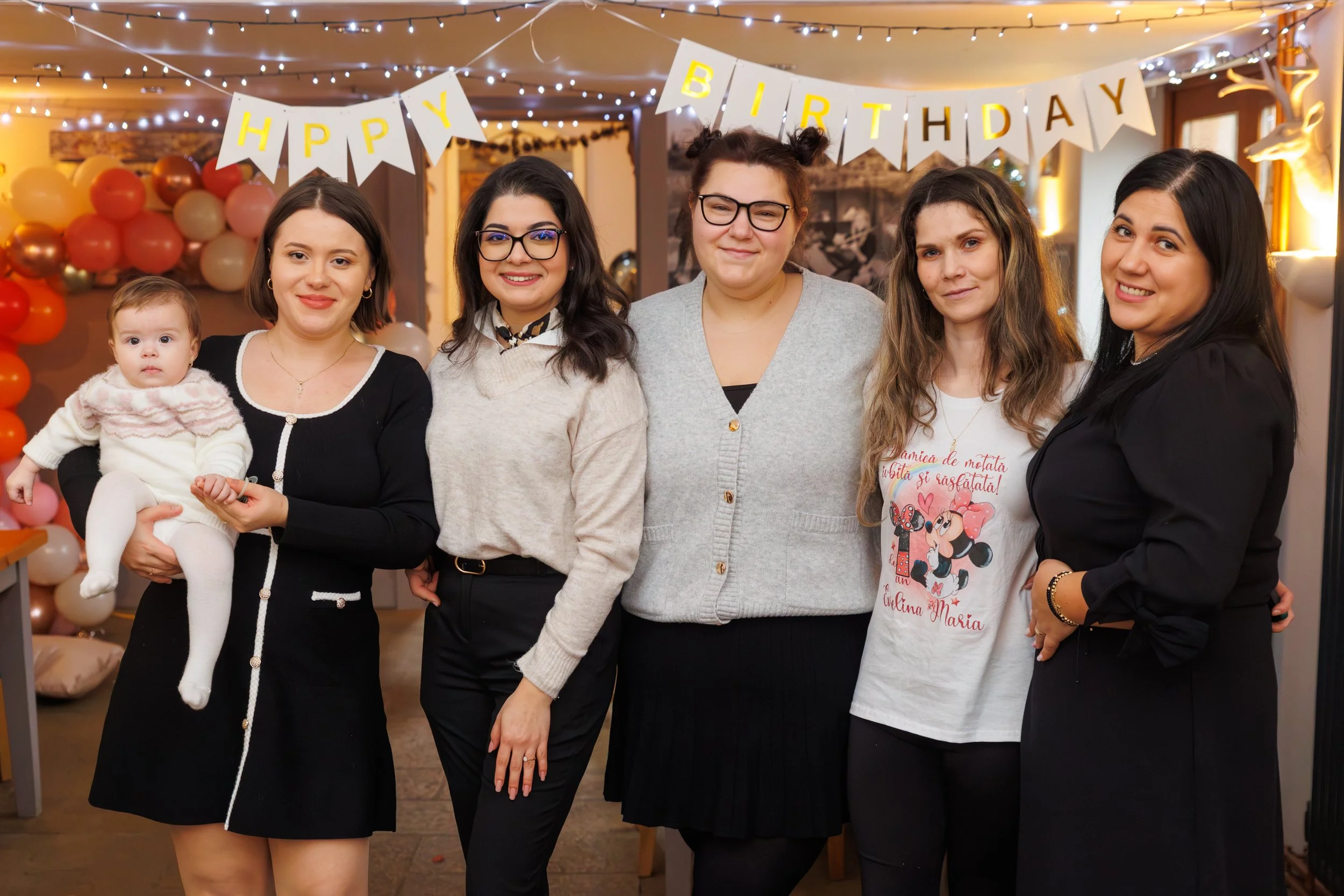 A group of women and a baby at a birthday celebration in a decorated room with balloons and fairy lights. There is a 'Happy Birthday' banner hanging above them.