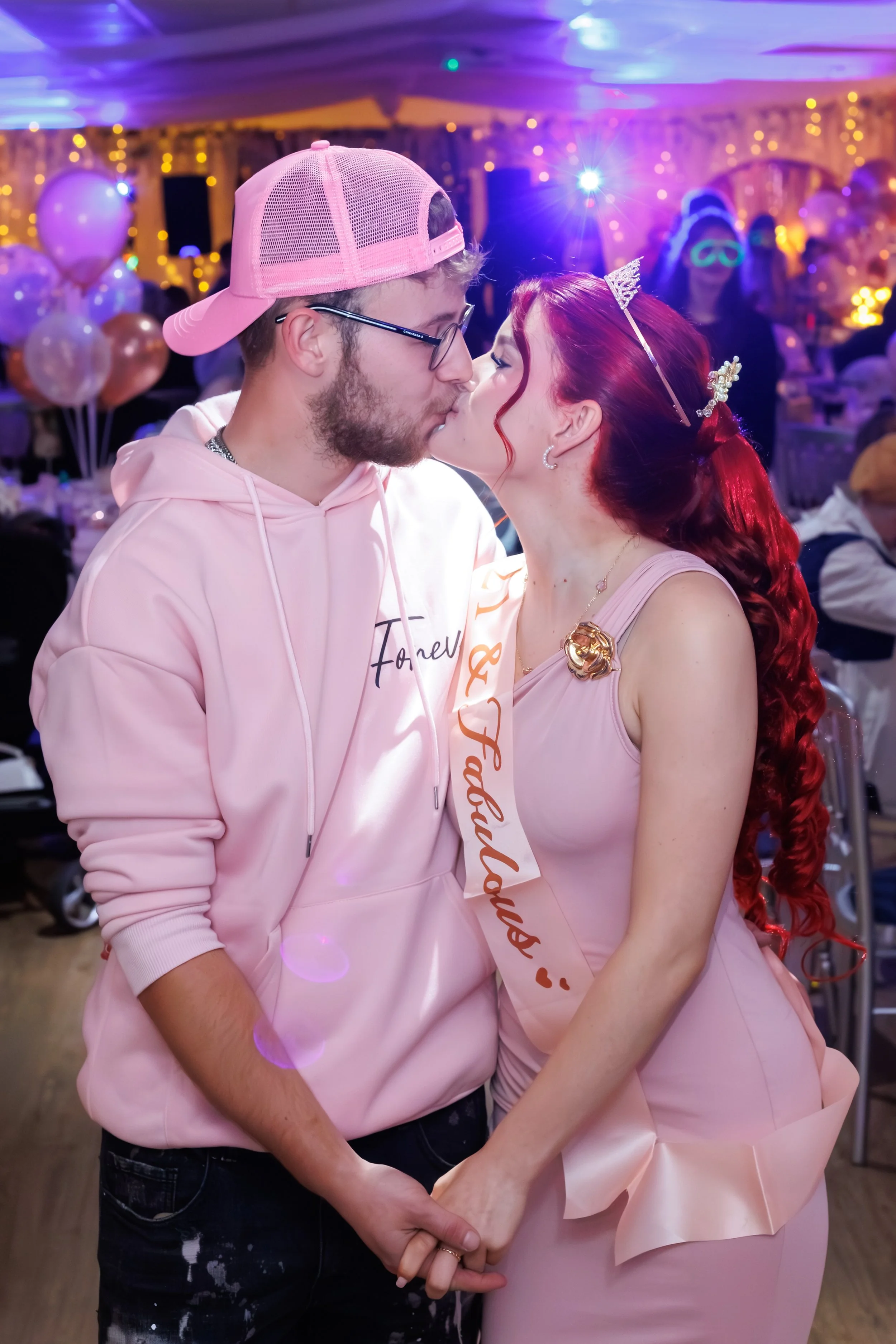 A couple kissing at a celebration or party, with festive balloons and colorful lighting in the background. The woman is wearing a pink dress, a tiara, and a sash, while the man is dressed casually with a pink hoodie and a pink cap.