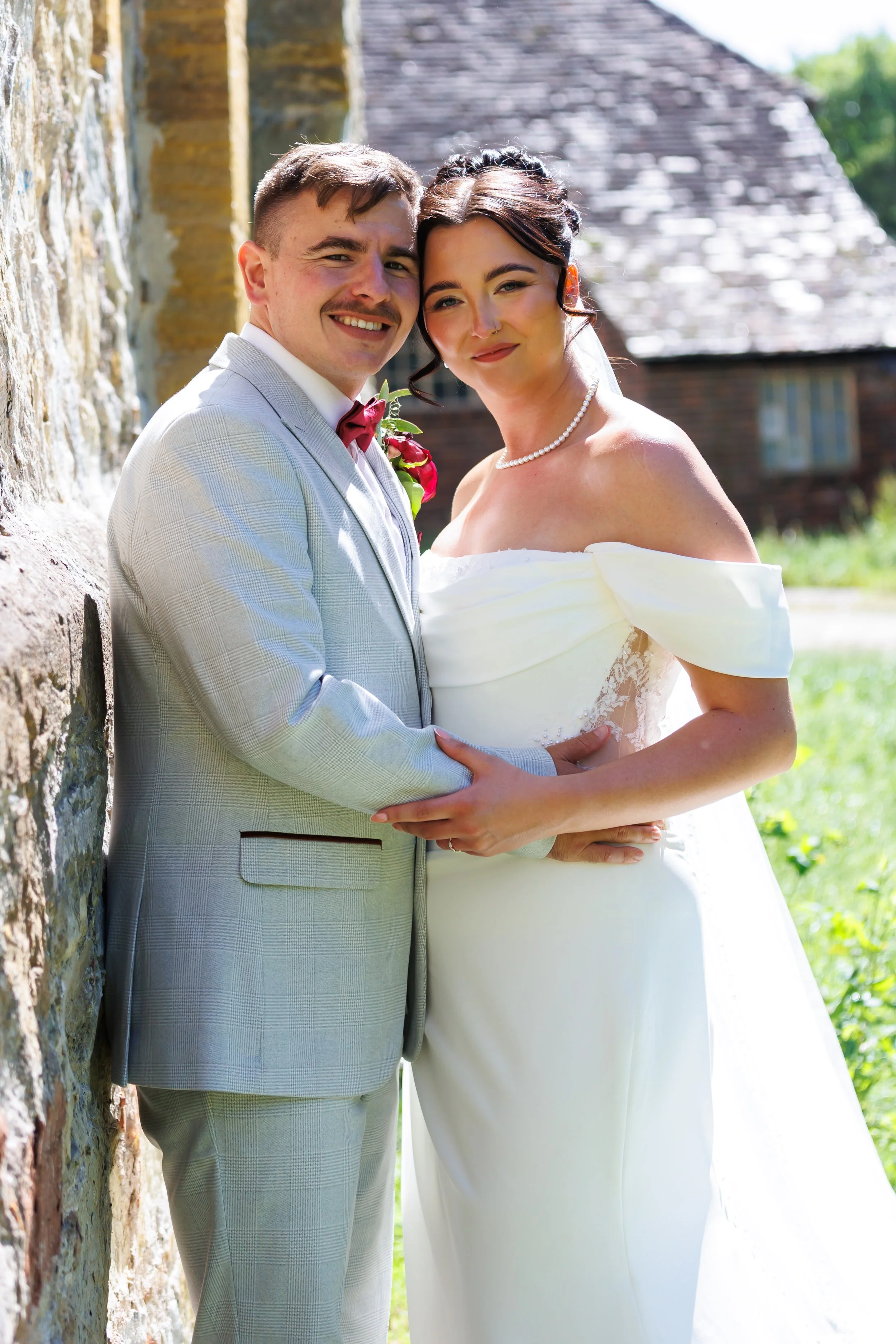 A smiling bride and groom dressed in wedding attire holding hands outdoors, standing close together with a stone wall and a rustic building in the background.
