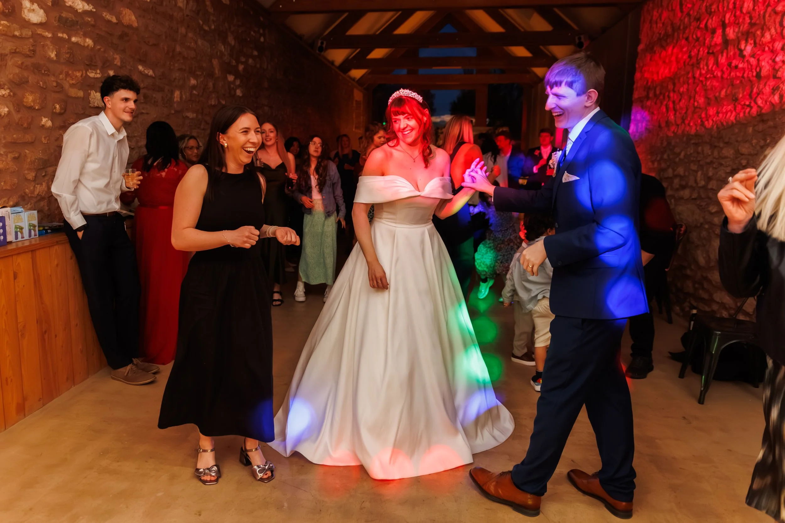 A bride in a white wedding dress with a tiara, dancing and smiling with guests at a wedding reception. Guests are gathered around in a decorated indoor venue with warm lighting.