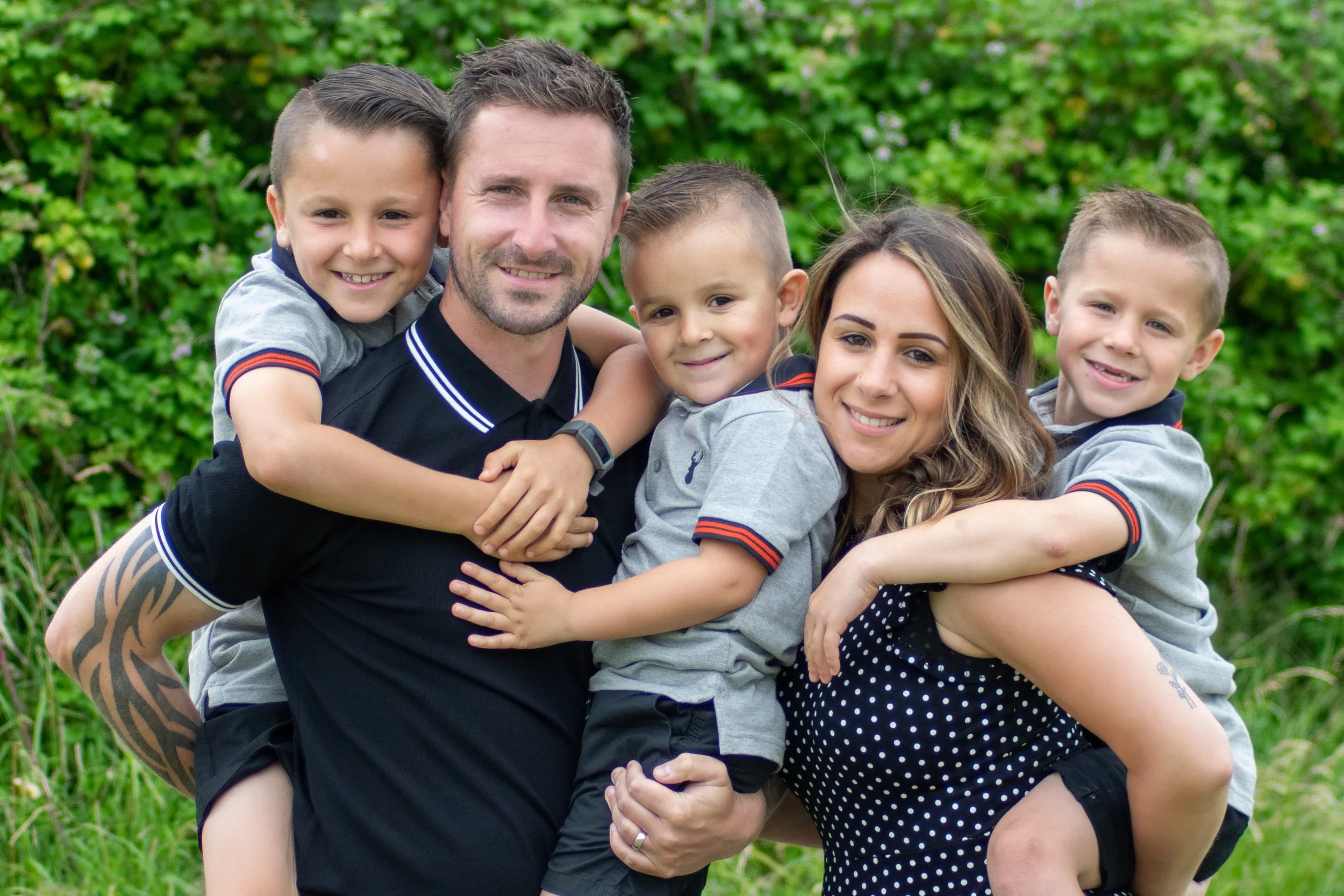 A family of five smiling and hugging outdoors in front of green foliage.