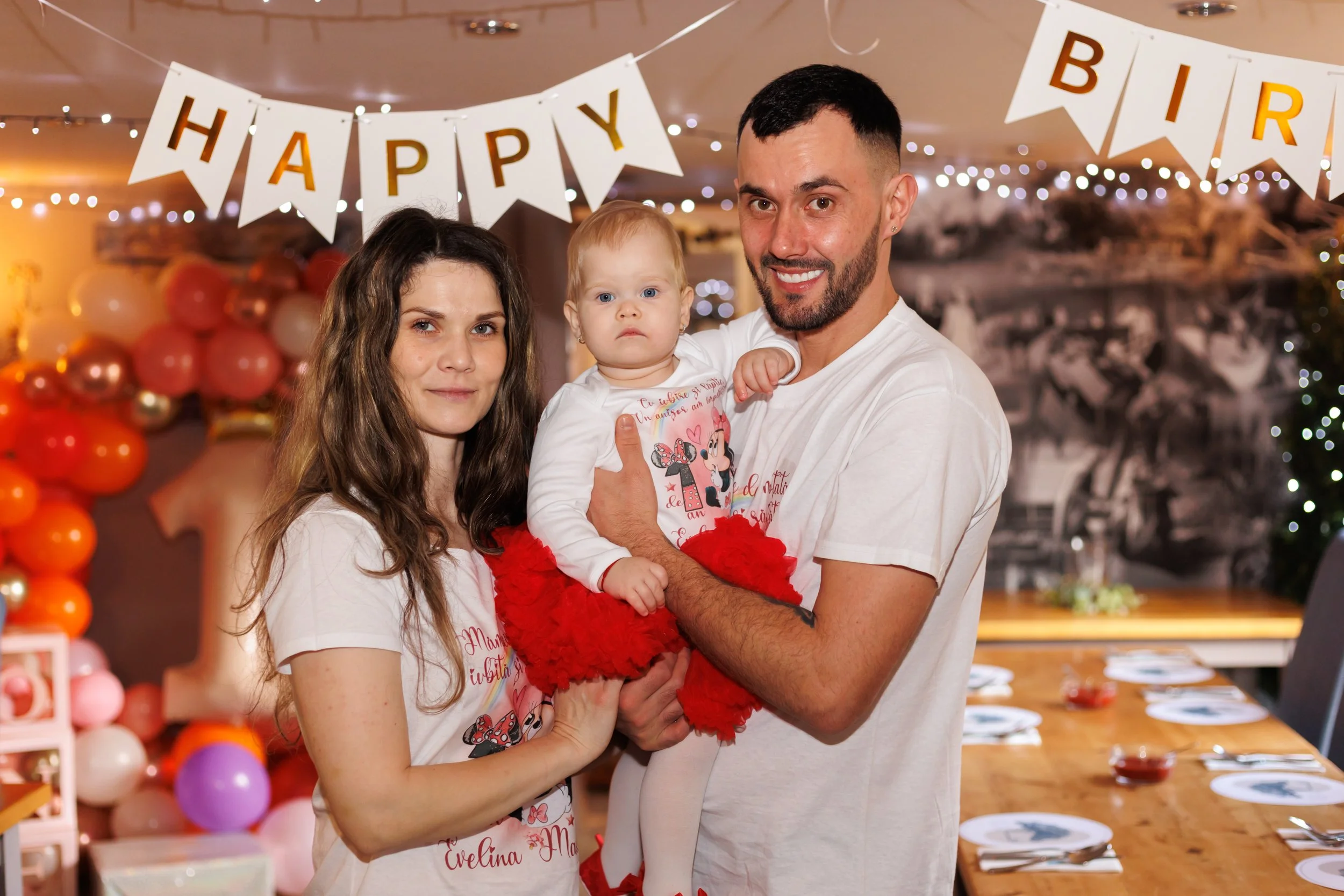 A family of three celebrating a birthday indoors, with decorations including a banner that says 'HAPPY BIRTHDAY' and balloons in the background. The group includes a woman with long brown hair, a man with a short beard, and a young girl with blonde h